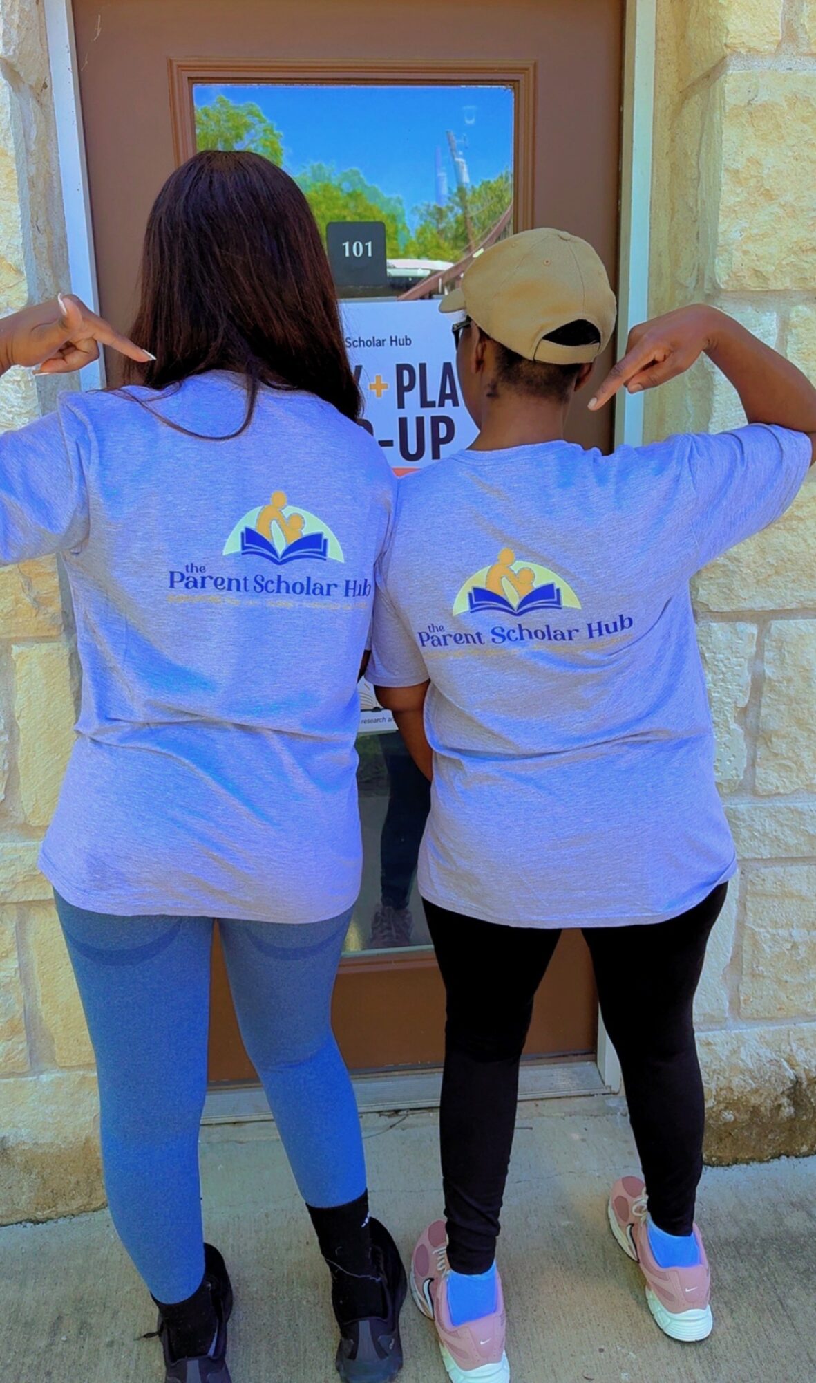 Two children in matching blue shirts with a logo, standing outside, flexing their arms, facing a window with trees visible inside.