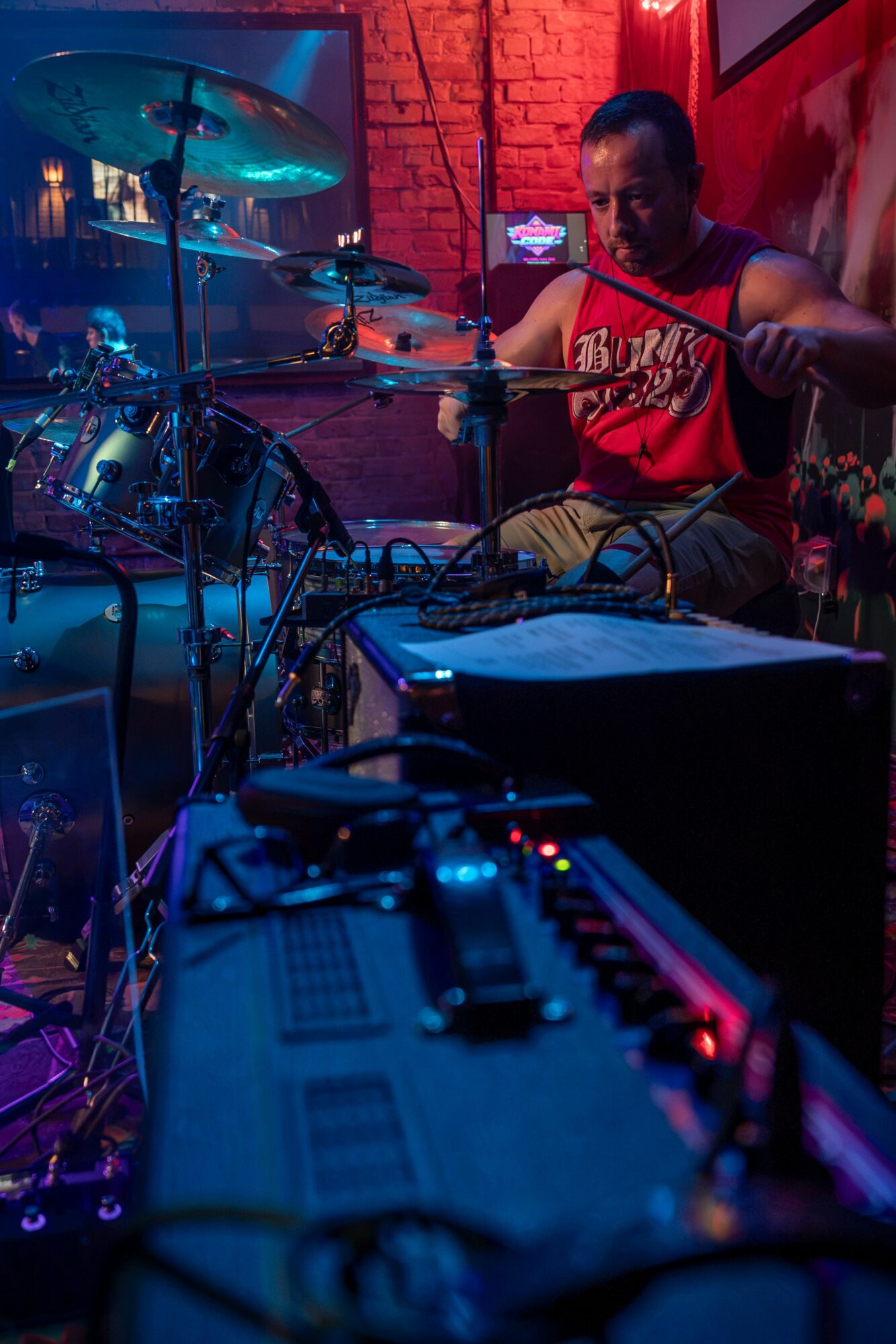 Musician playing drums in a dimly lit venue with colorful lighting and a brick wall background.