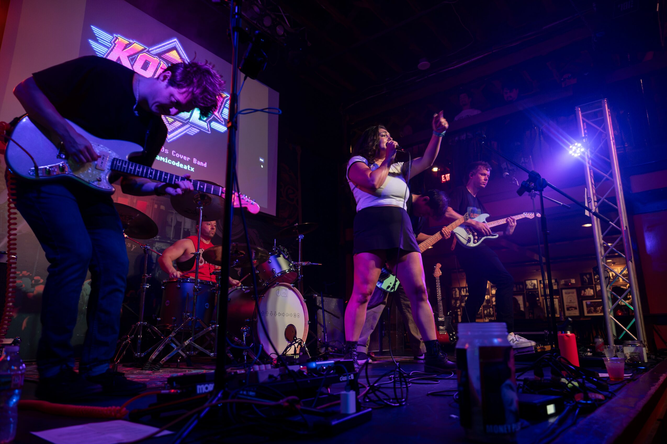 Band performs on stage with a singer, guitarist, bassist, and drummer, colorful stage lighting, and a screen in the background.