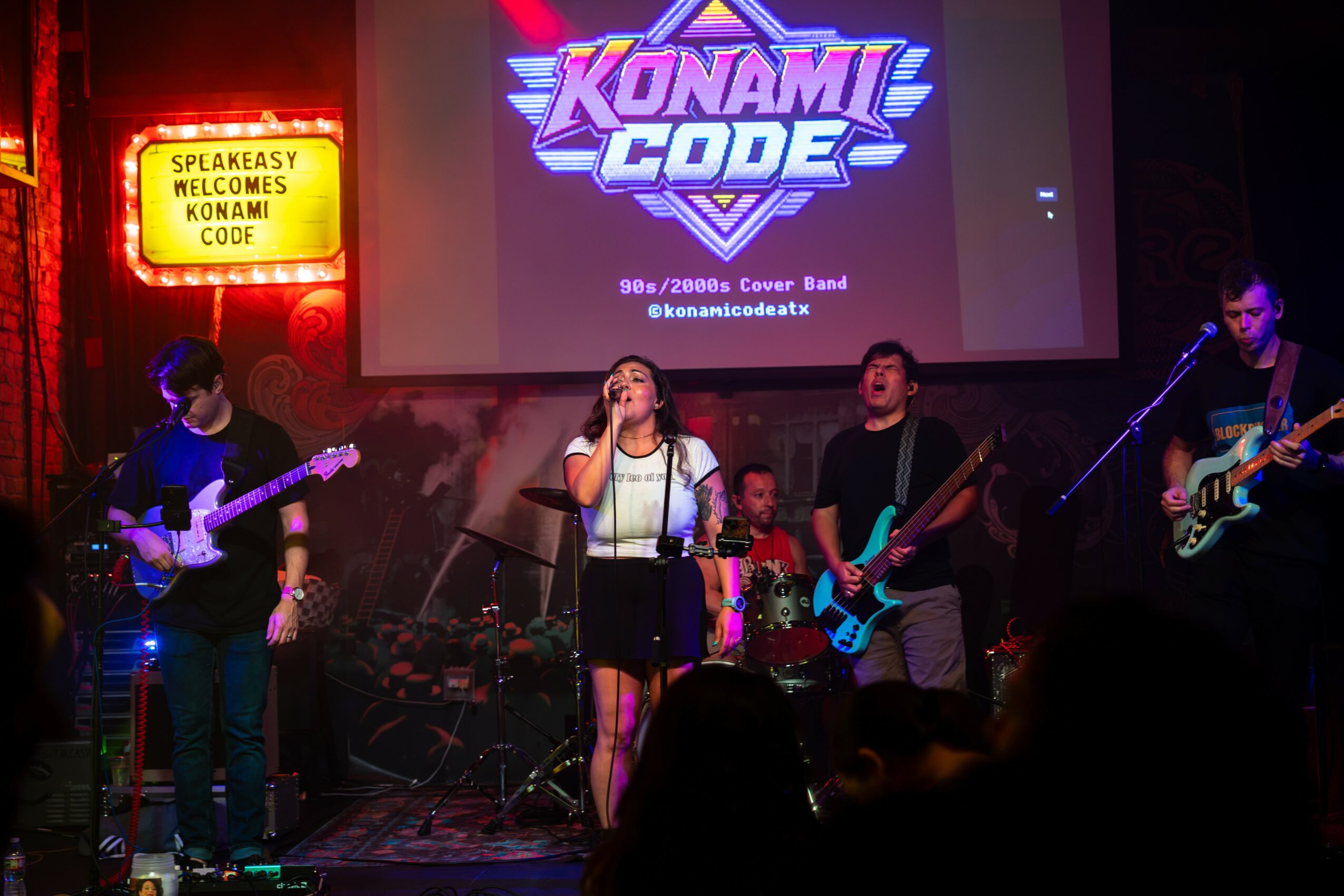 Band performs on stage with a large screen displaying 'Konami Code' behind them, audience visible in foreground.