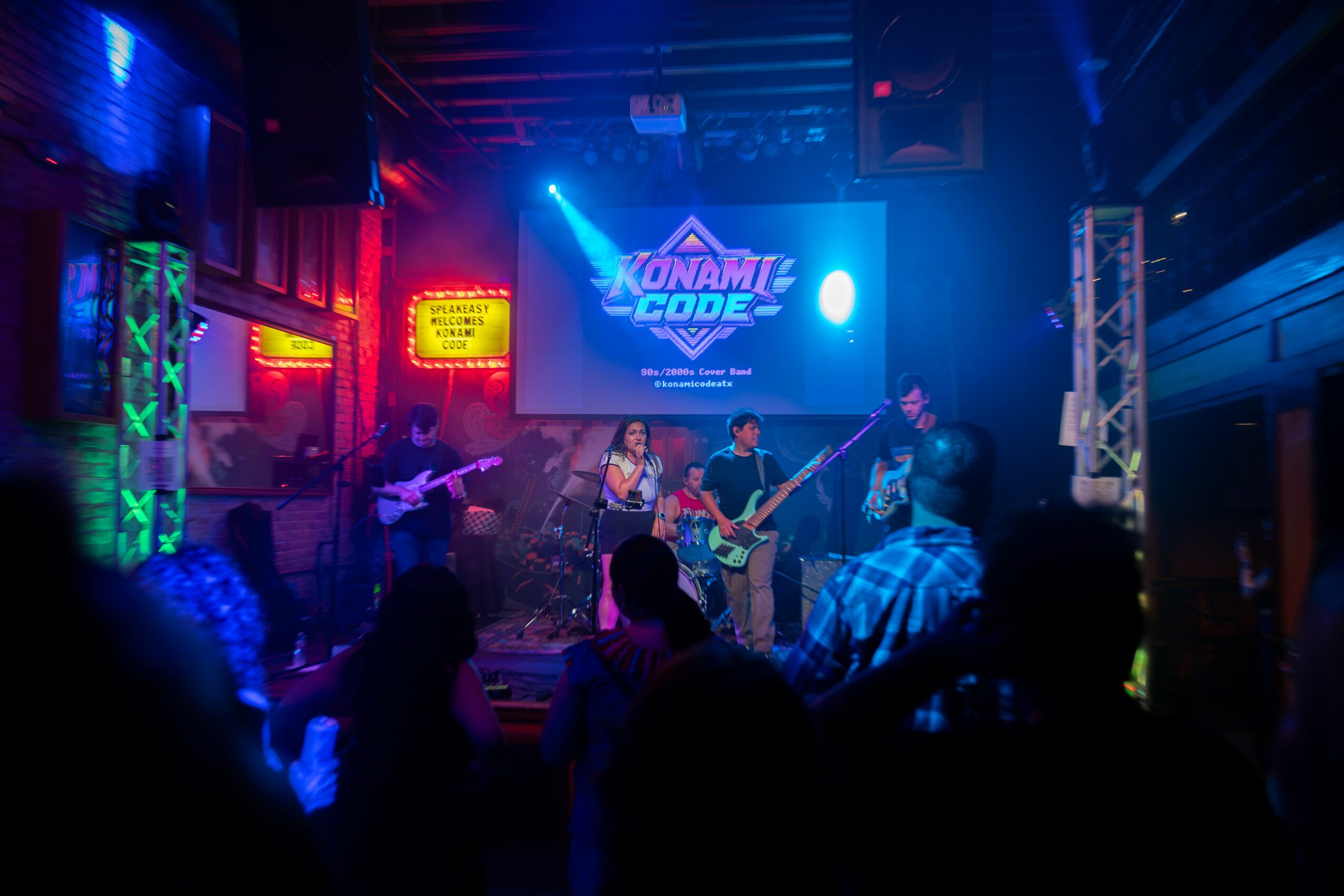 Band performs on stage with colorful lights and a large screen displaying 'KONAMIE CODE' in a lively venue.