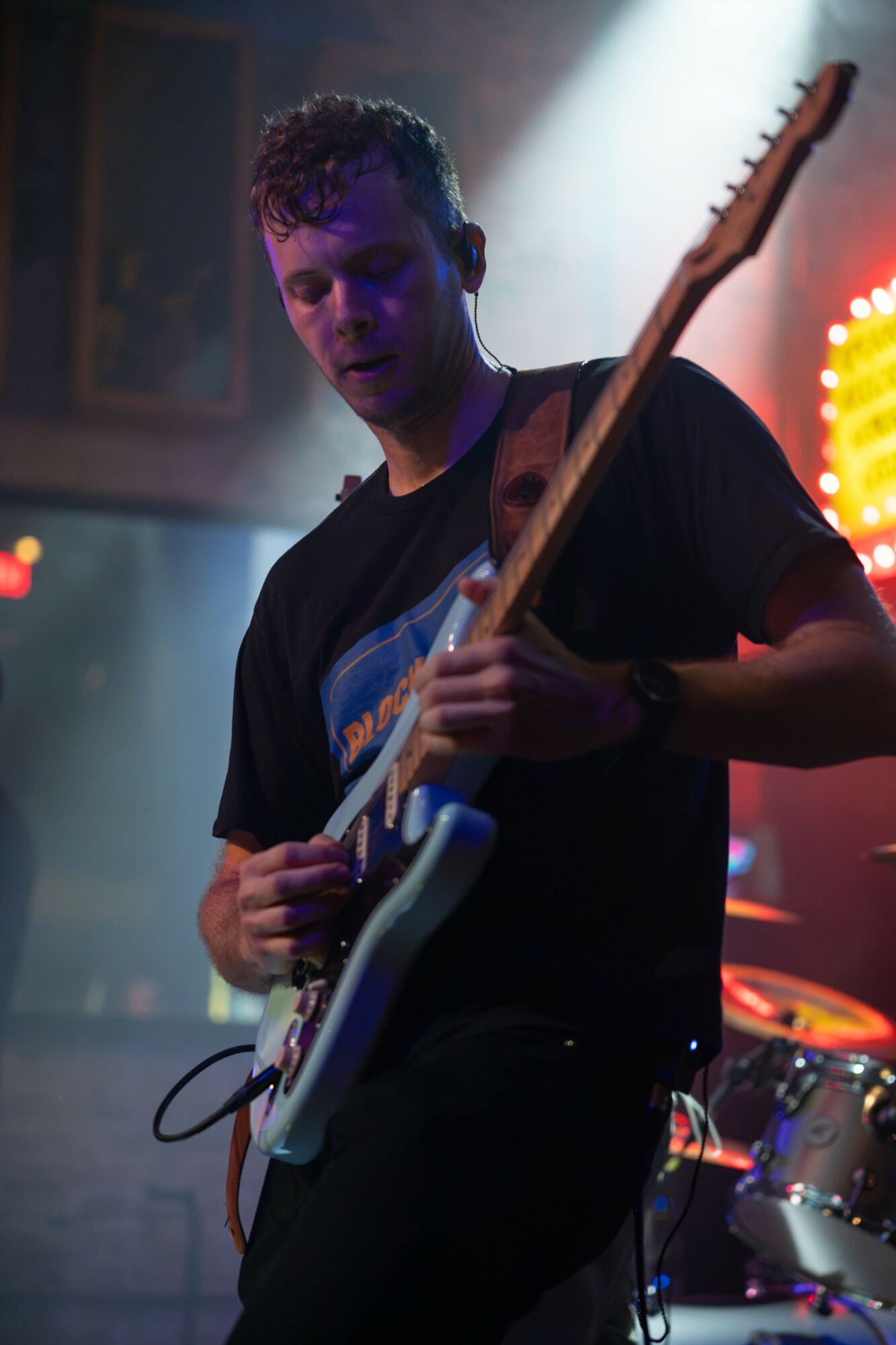 Young man playing electric guitar on stage with colorful lights in background.