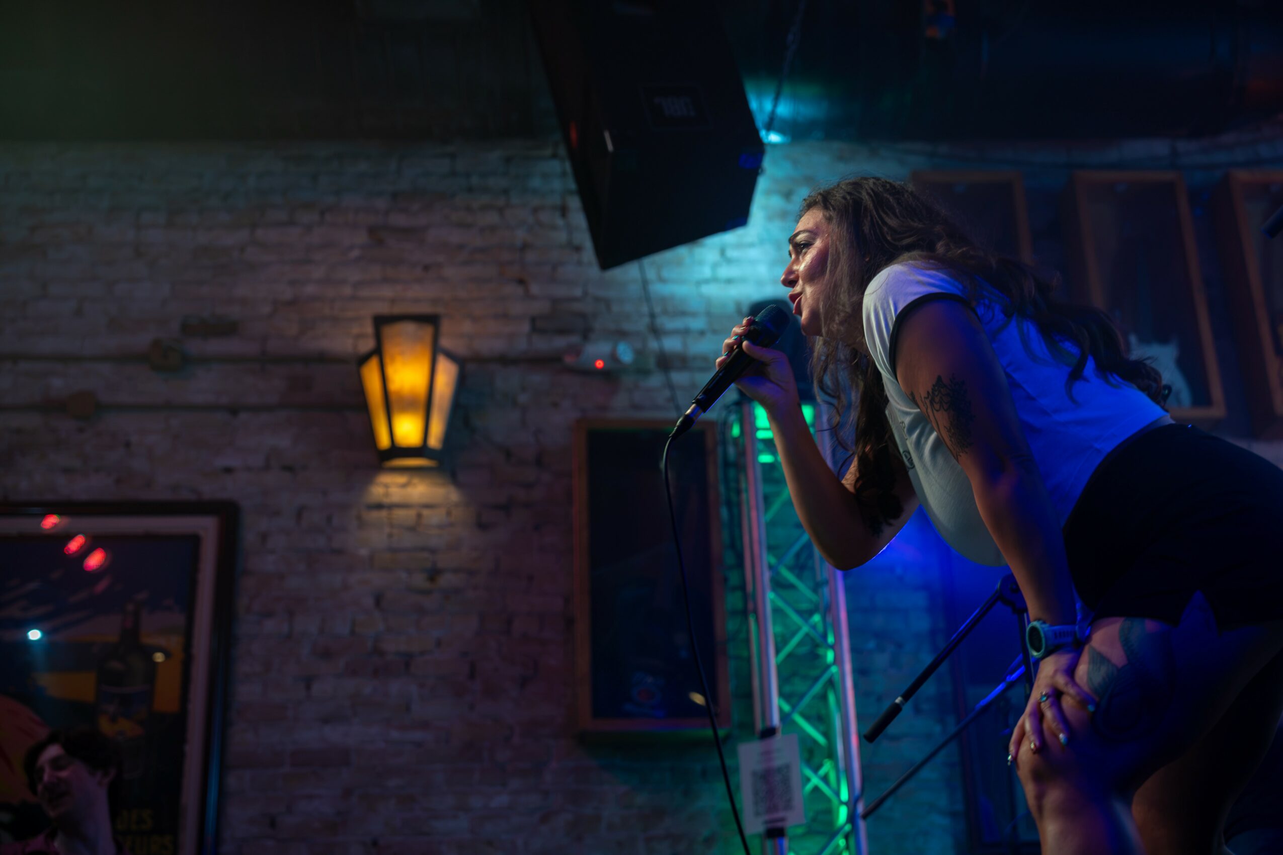 A woman sings into a microphone on stage with colorful lighting, brick wall background, and a wall-mounted lamp.