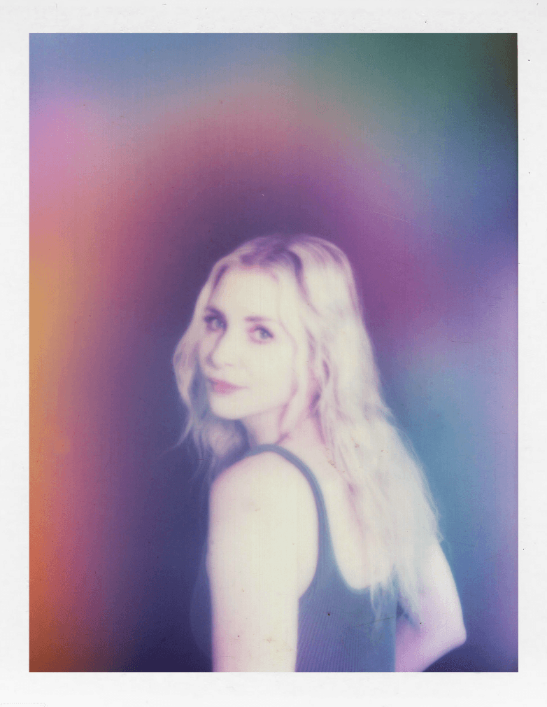 A woman with long, wavy blonde hair looks over her shoulder, smiling, with a colorful, blurred background.