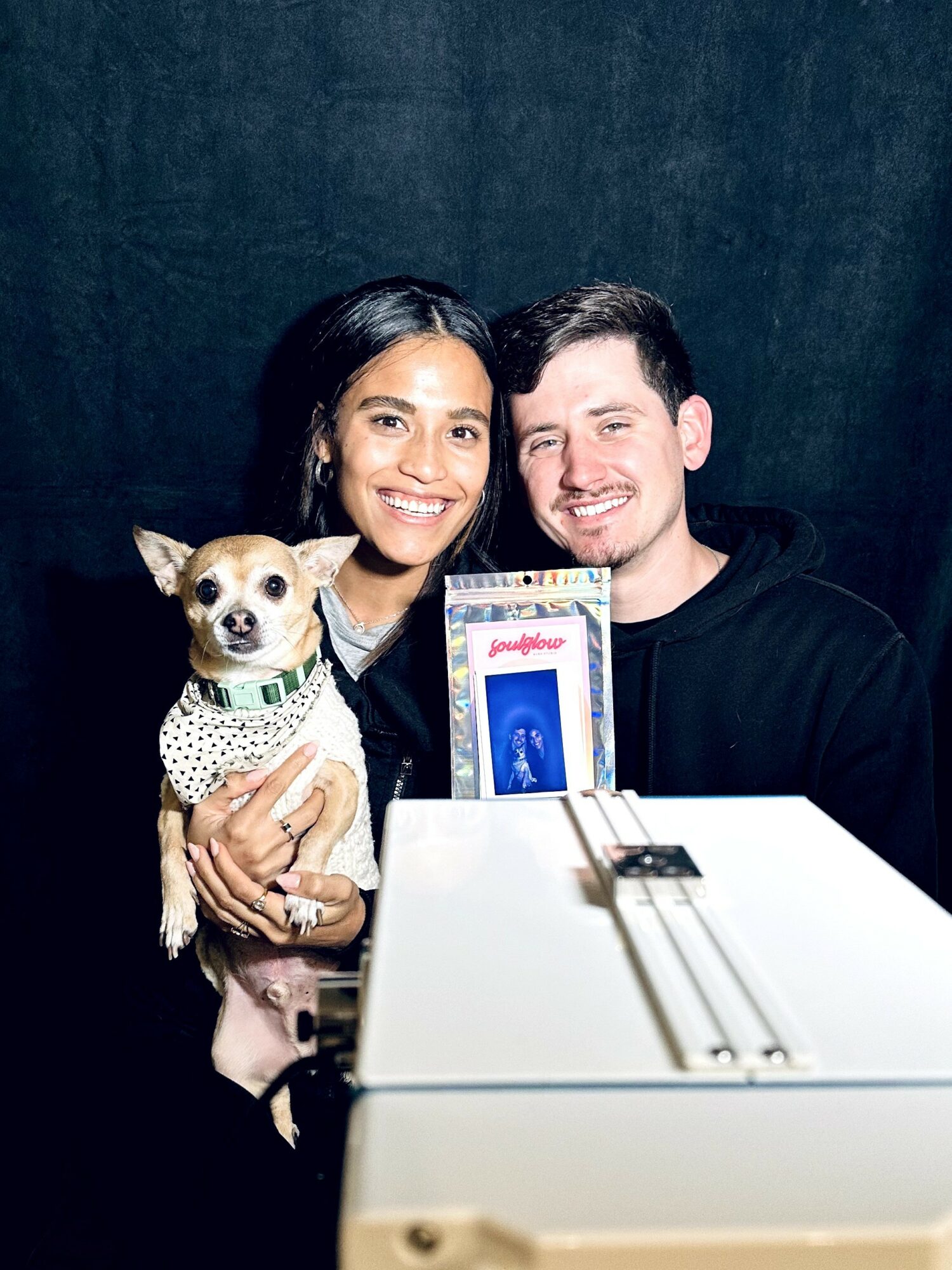 Two people smiling with a small dog, holding a phone, in front of a dark background.