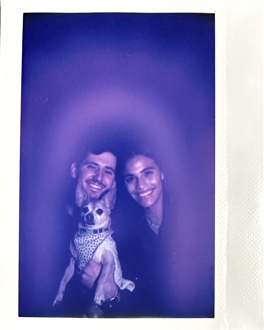 Two people smiling with a small dog between them, dark background, photo has a blue tint.