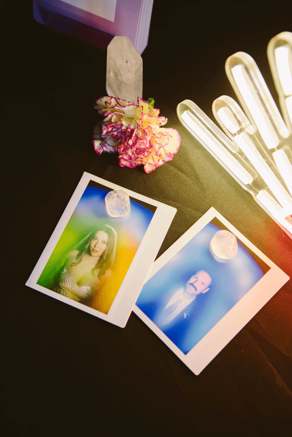 Two Polaroid photos on a dark surface, one showing a woman and the other a person, with a crystal and flowers nearby.
