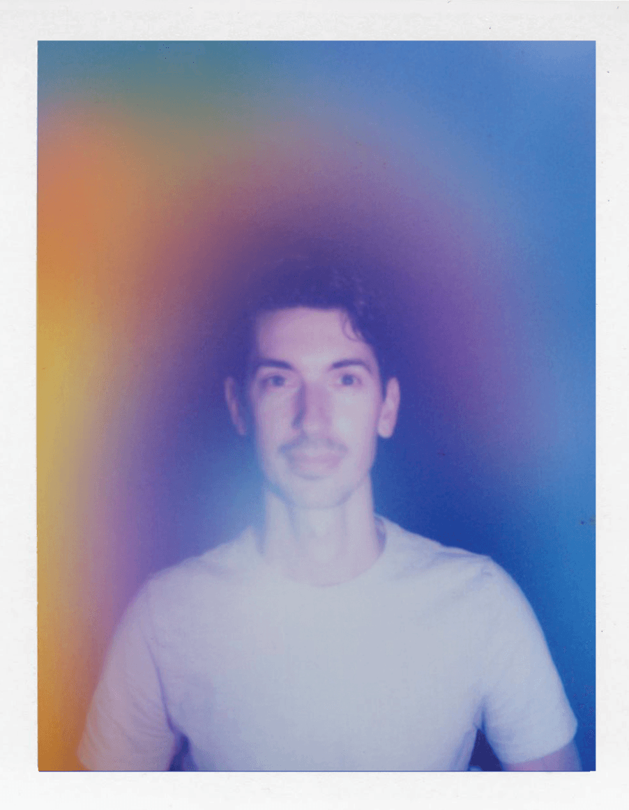 Young man with dark hair and a light-colored shirt, smiling, against a colorful, blurred background.