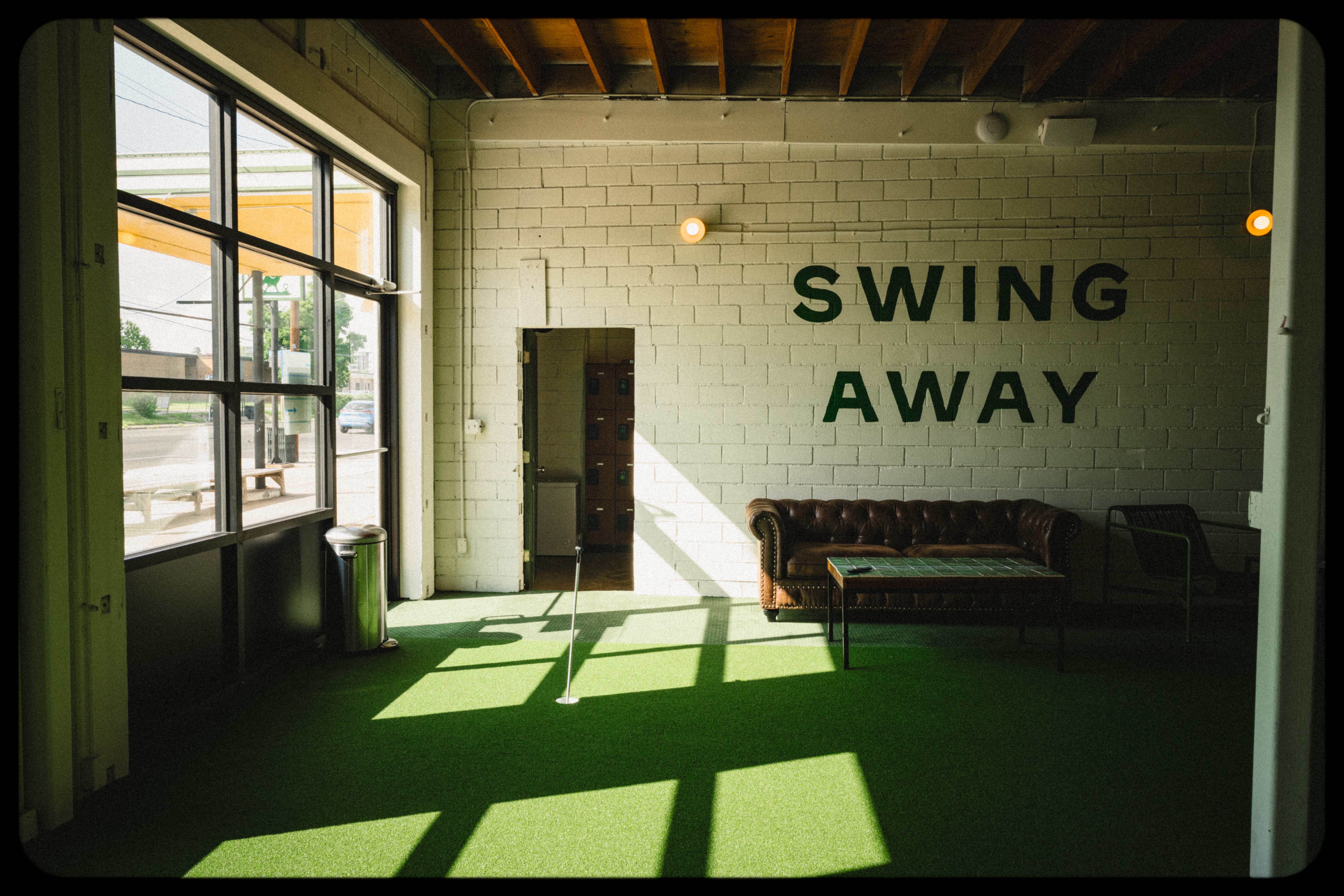 Sunlight casts shadows on a green floor inside a room with a white brick wall, a sofa, and the words 'SWING AWAY' on the wall.