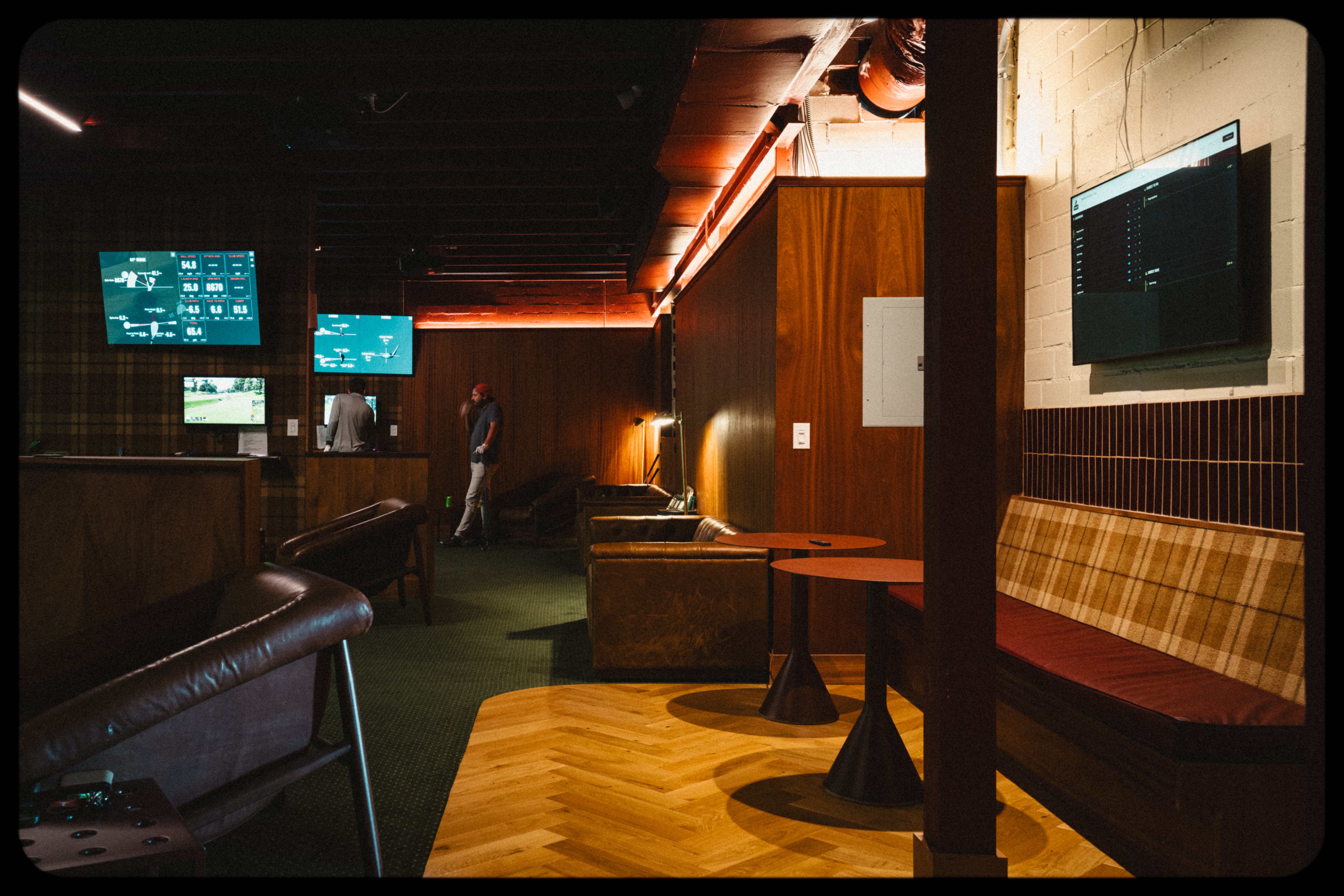 Interior of a dimly lit lounge with seating, tables, and screens on the walls, and a person standing in the background.