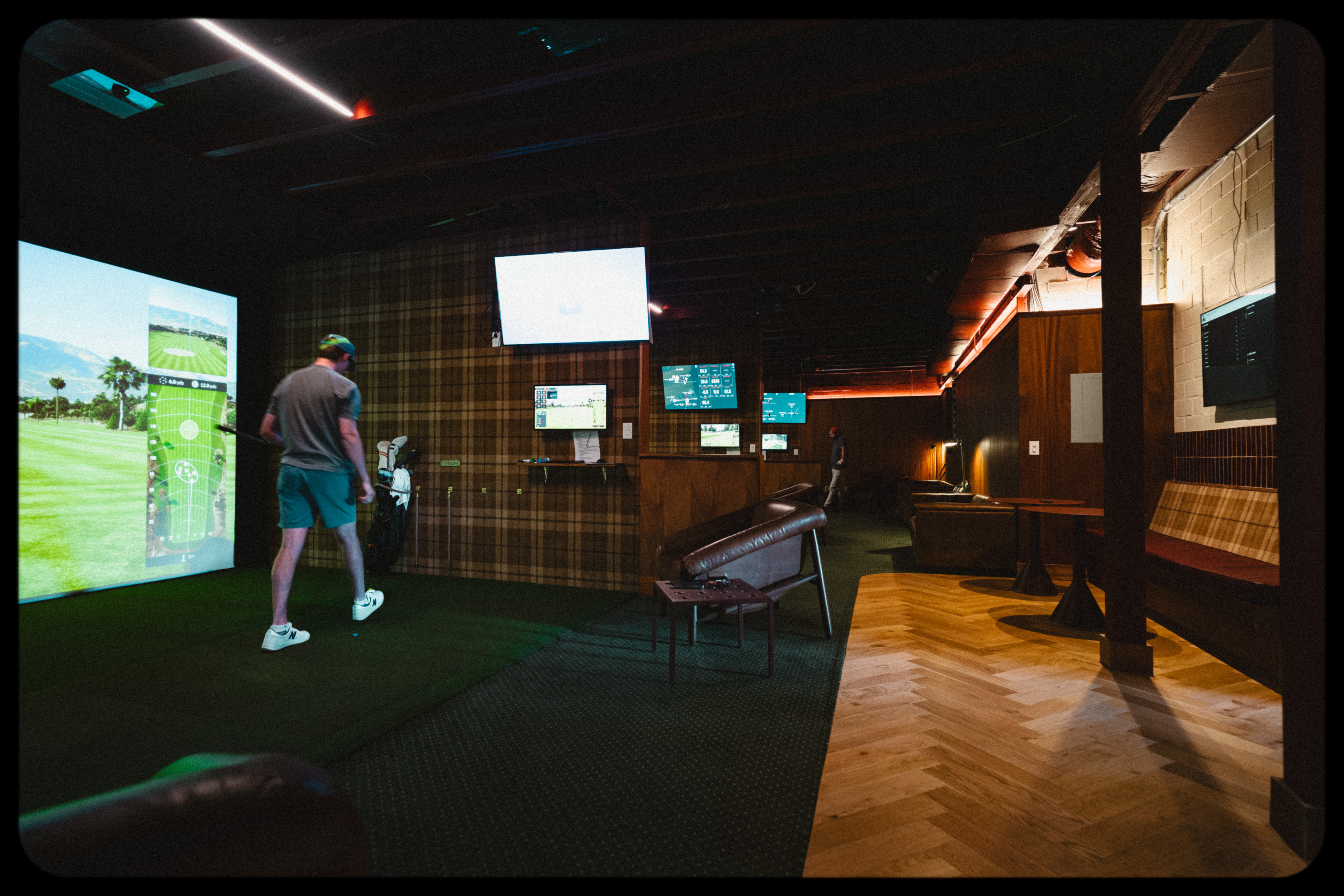 Indoor golf simulation room with a person playing, multiple screens, and seating area, dim lighting, wooden flooring, and modern decor.