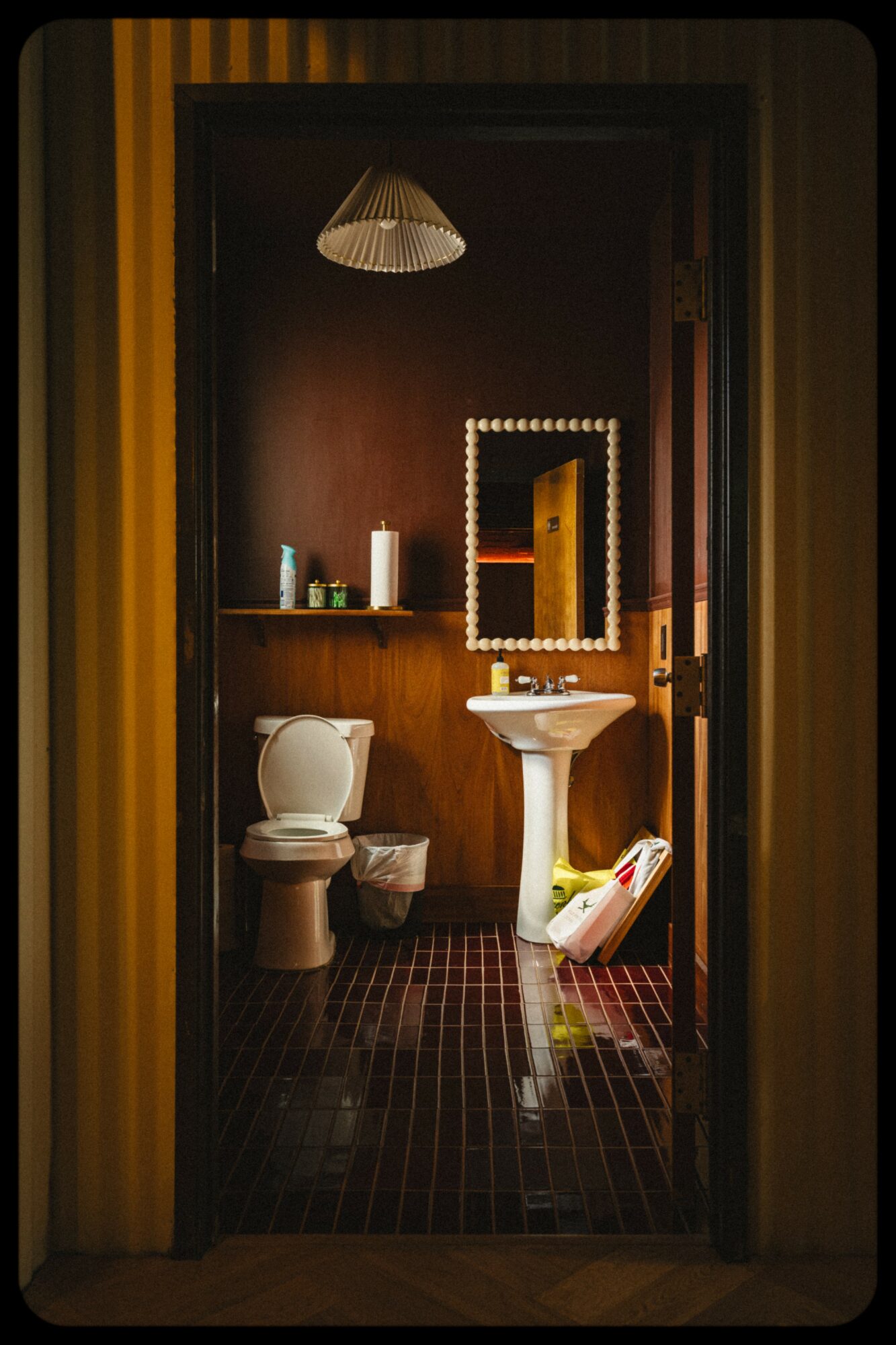 Bathroom with a toilet, pedestal sink, mirror, and shelf, viewed through a doorway with yellow and brown walls.