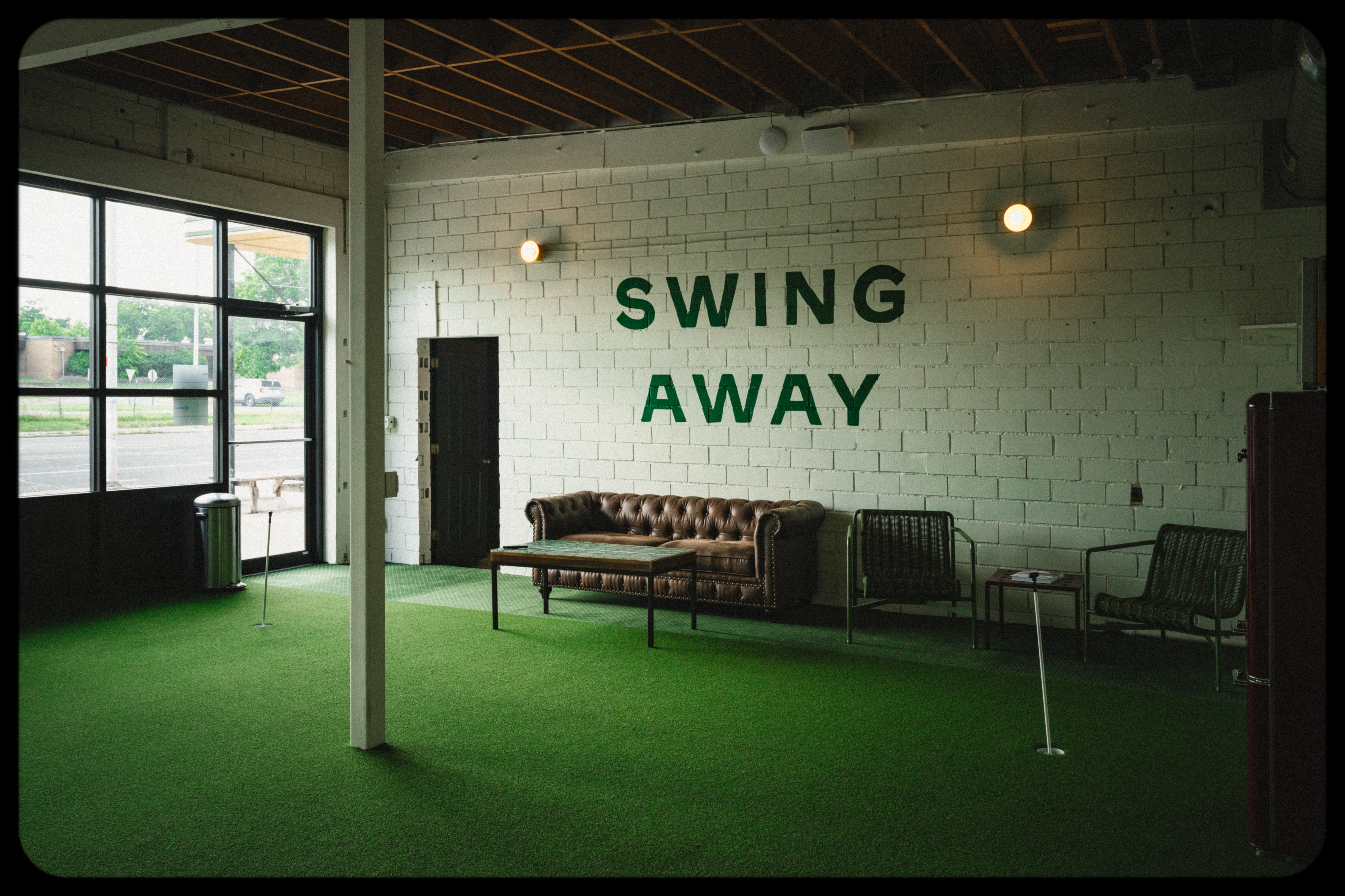 Indoor space with green carpet, a brown sofa, chairs, and a wall with 'SWING AWAY' text, large windows on the left.