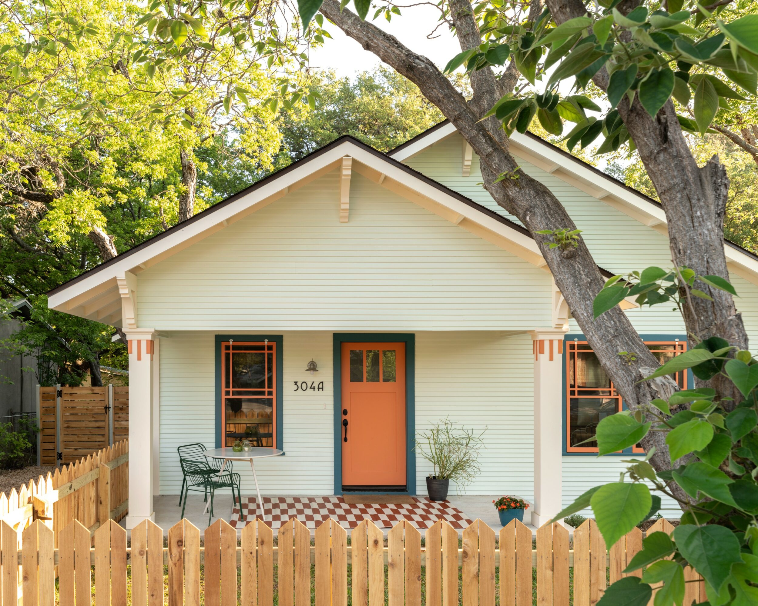 Front view of a small house with a orange door, two windows, a porch, and a wooden fence, surrounded by trees.