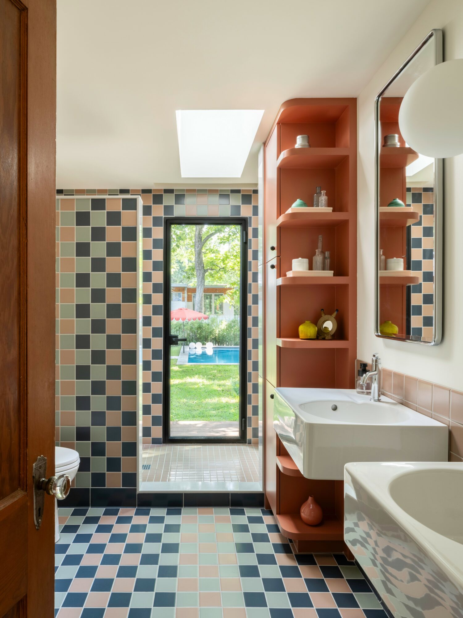 Bathroom with patterned tile floor, open door to outside, pink shelving, white sink, mirror, and bathtub, natural light from skylight.