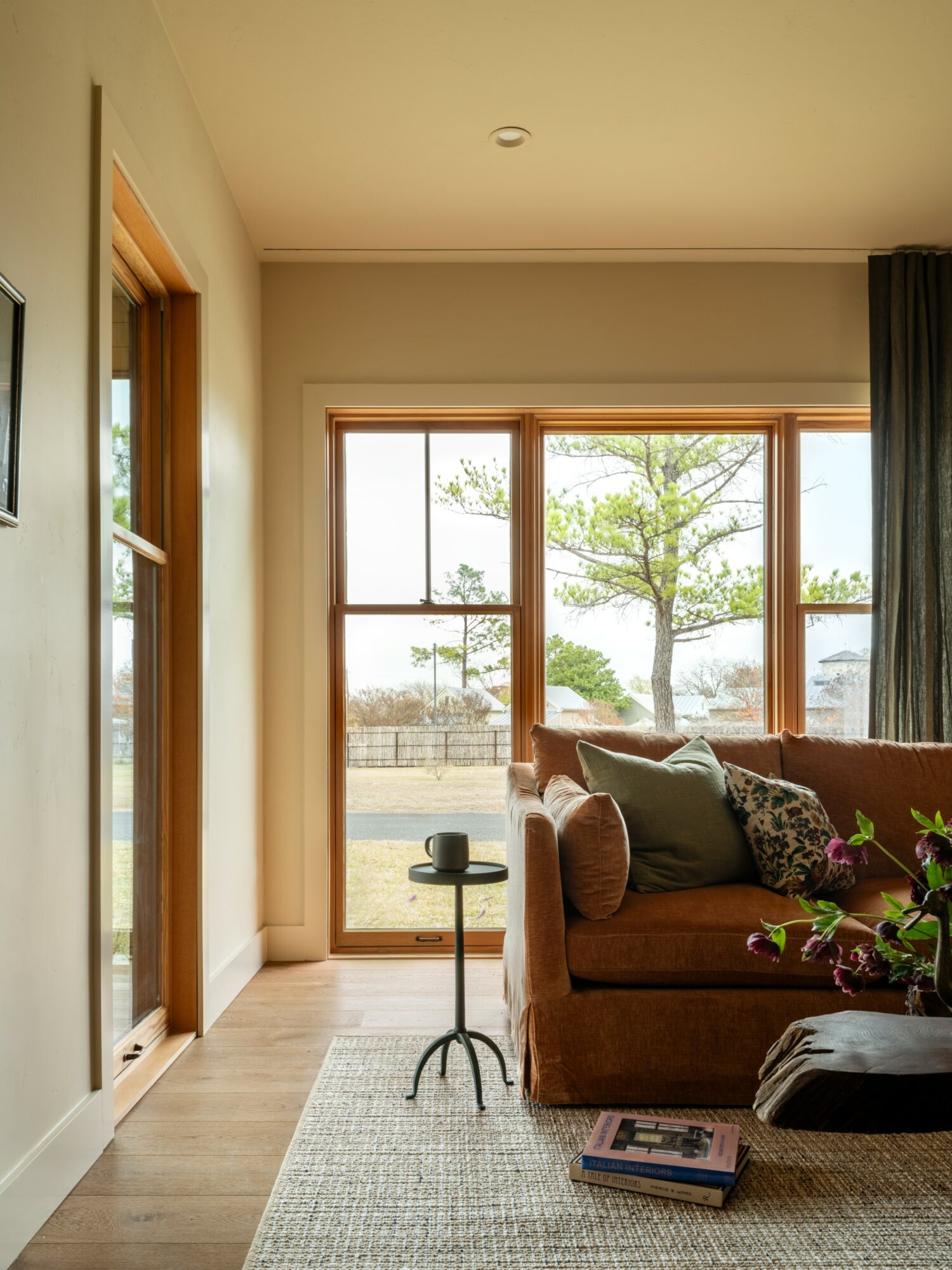 Living room with large window showing outdoor trees, brown sofa with pillows, side table, and books on the floor.