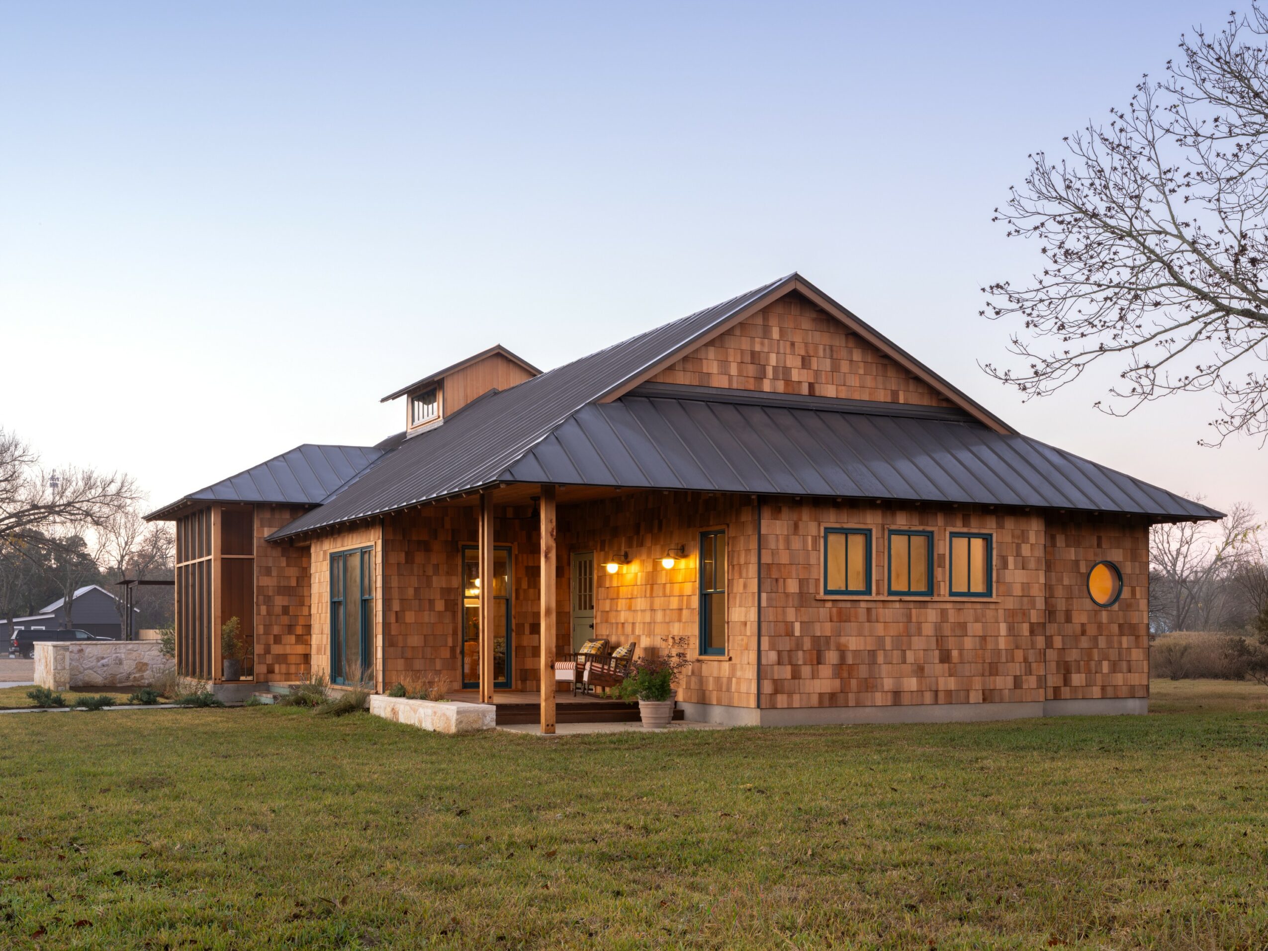 A house with a metal roof and wooden exterior, surrounded by a grassy yard and trees, during sunset.