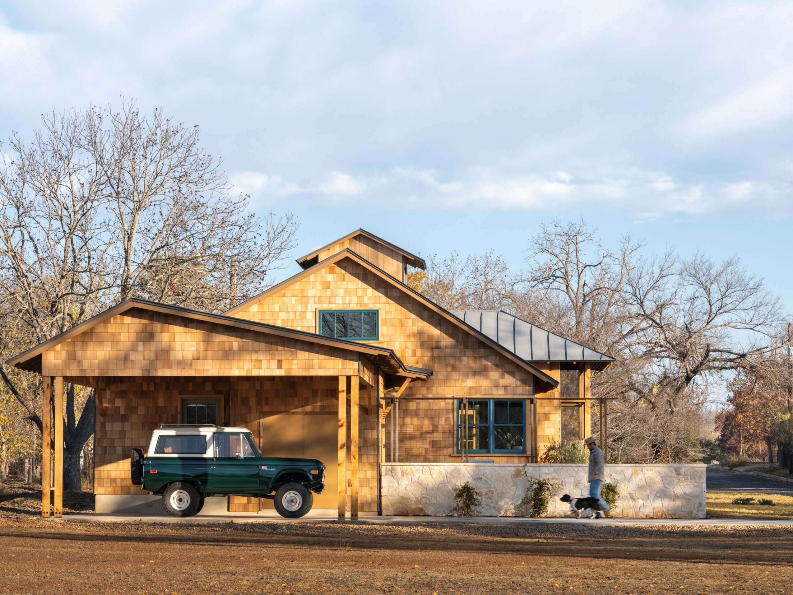 A wooden house with a car parked in front, trees in the background, and a partly cloudy sky.