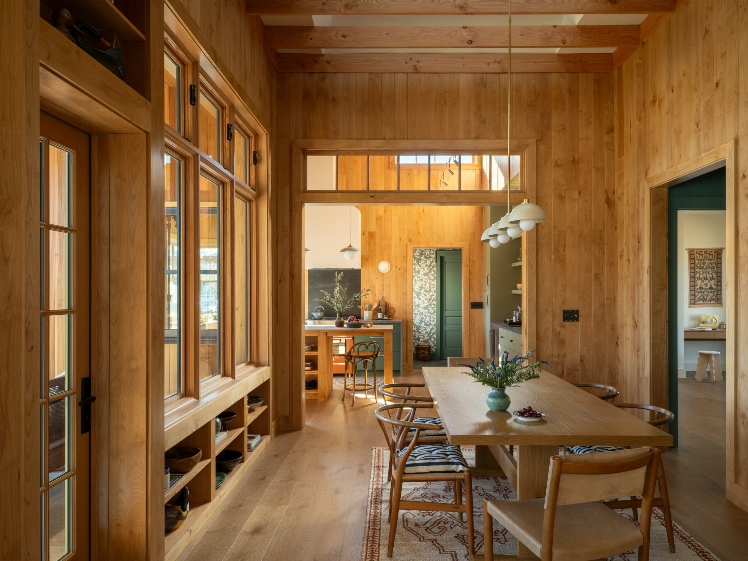 Wood-paneled dining room with a long table, chairs, and large windows, leading to a kitchen area in the background.