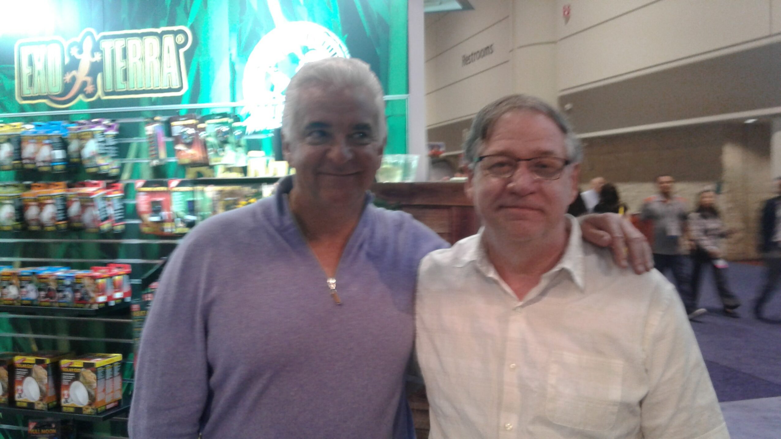 Two people standing together indoors, one with gray hair and the other with glasses, smiling, with a store in the background.