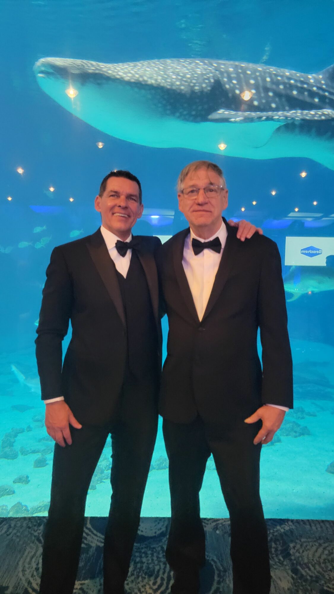Two men in formal suits standing in front of an aquatic-themed background with whales and water effects.