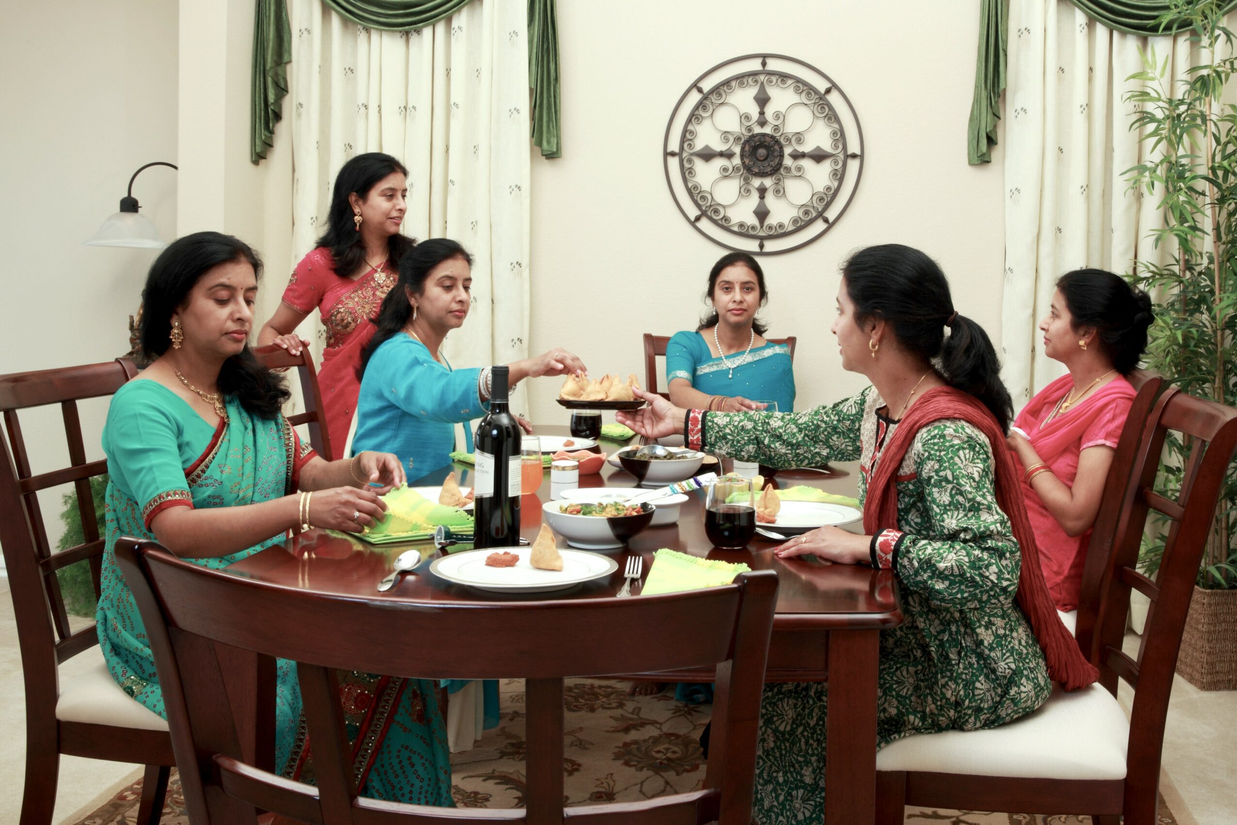 Seven women sit and stand around a dining table in a room with green curtains and a clock on the wall, sharing a meal.