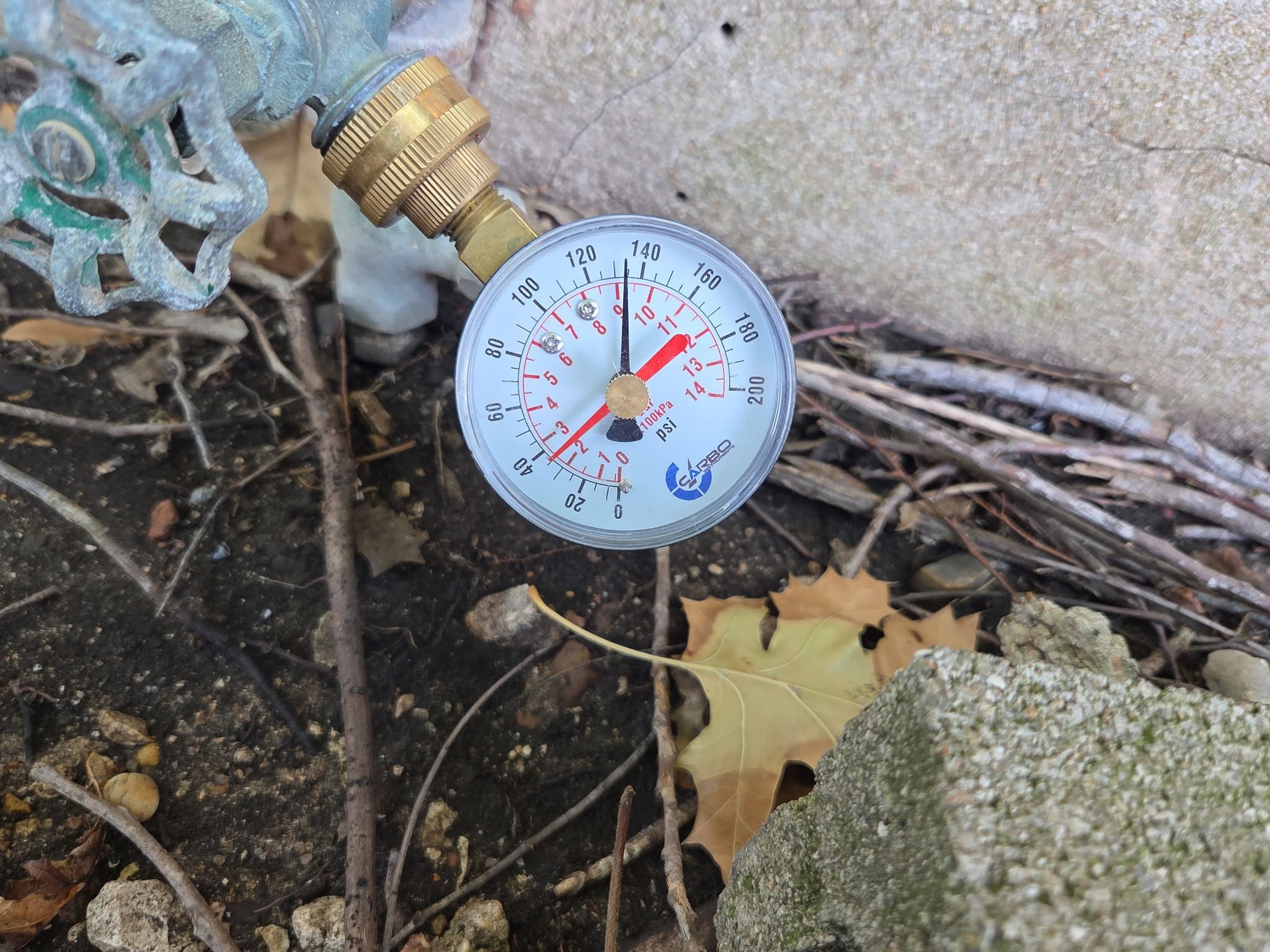 Pressure gauge attached to a pipe outdoors, with soil, leaves, and a concrete surface nearby.