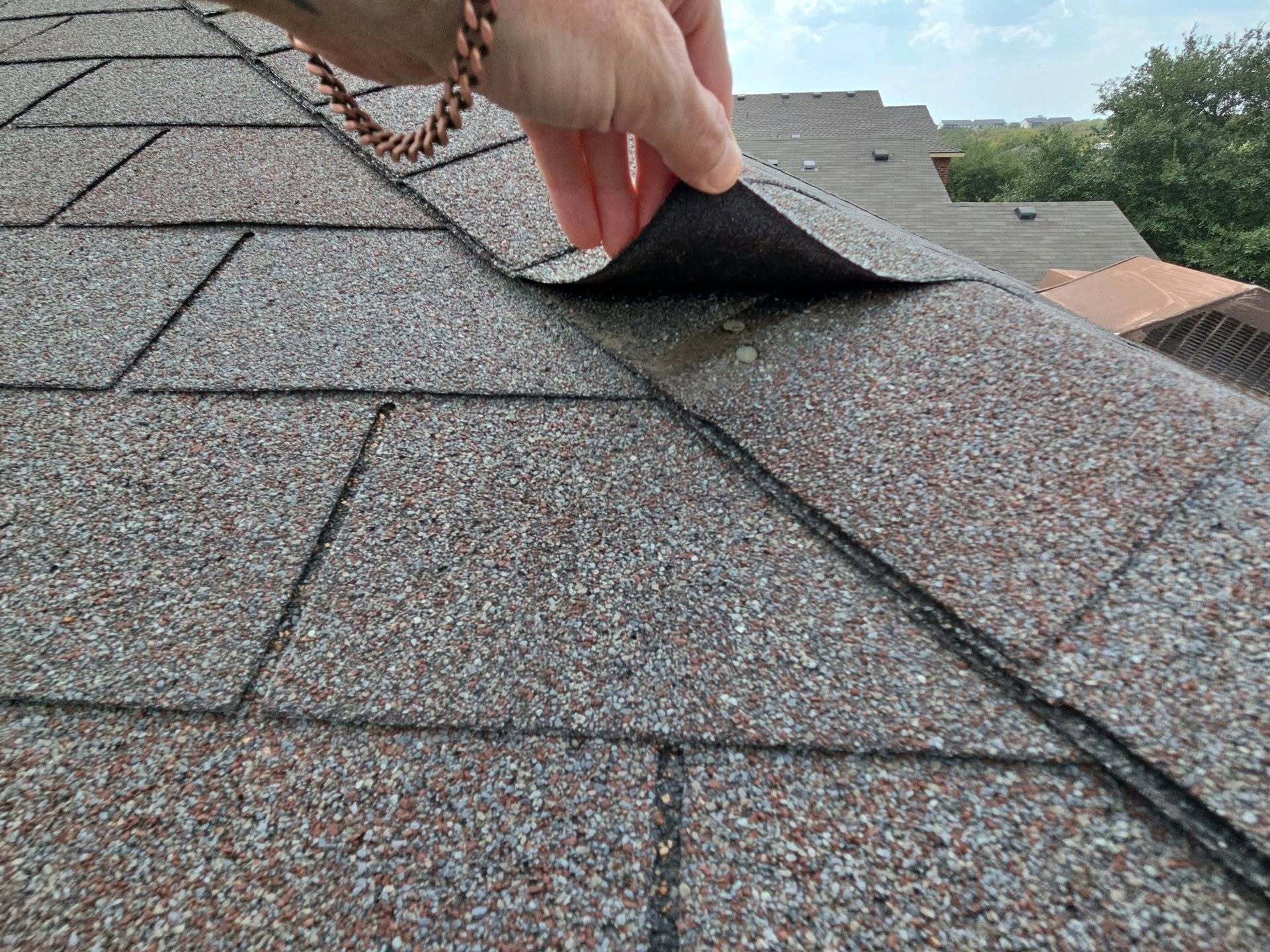 Person lifting shingle on roof with other shingles visible, sky and trees in background.