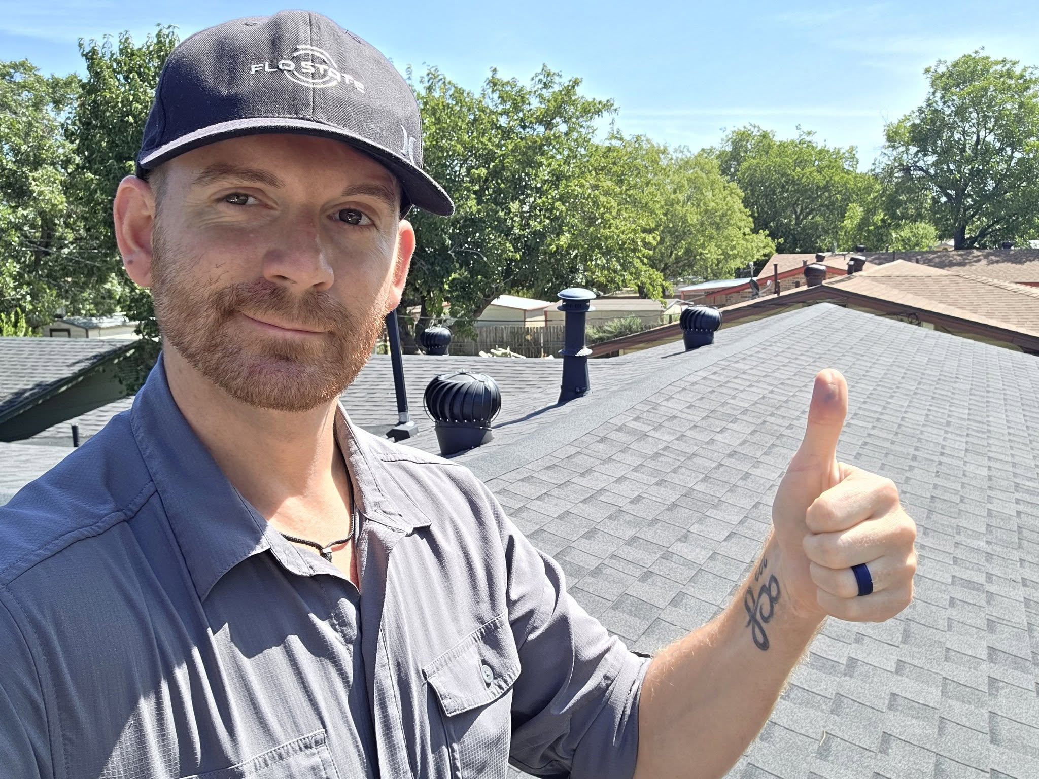 Man with beard and cap giving a thumbs-up on rooftop, trees and blue sky in background.