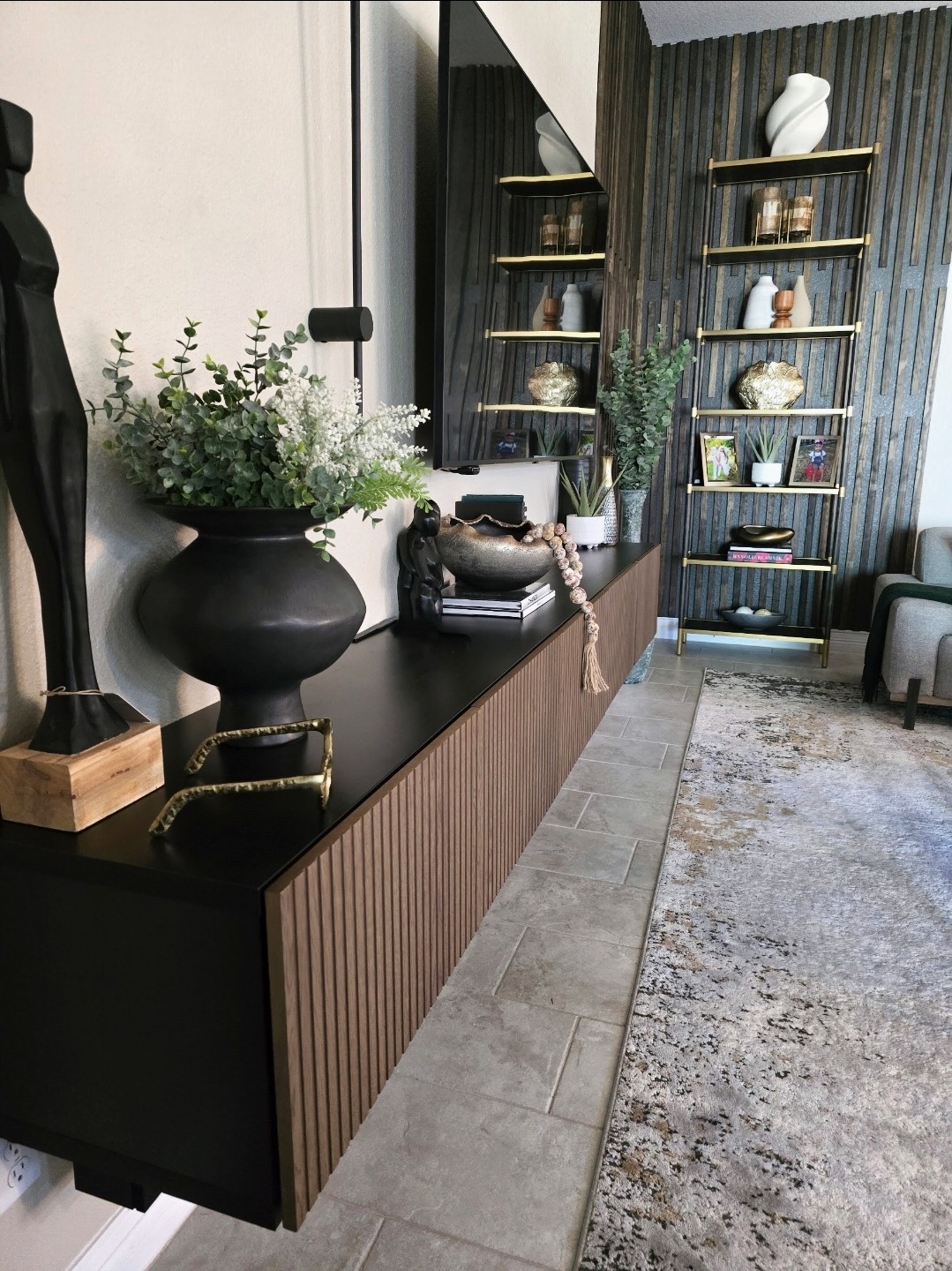 Interior room with black and wood sideboard, decorative vases, plants, and shelves with ornaments, rug, and seating area.