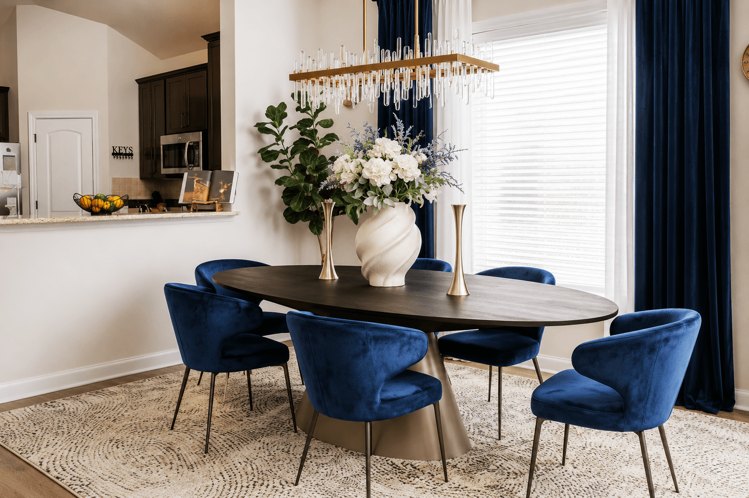 Dining area with a wooden table, five blue velvet chairs, a large white vase with flowers, and a chandelier, near a window with blue curtains.