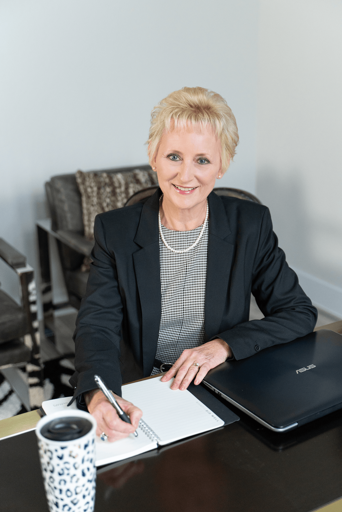 Smiling woman with short blonde hair, wearing a black blazer, sitting at a desk with a notebook, pen, laptop, and coffee mug.