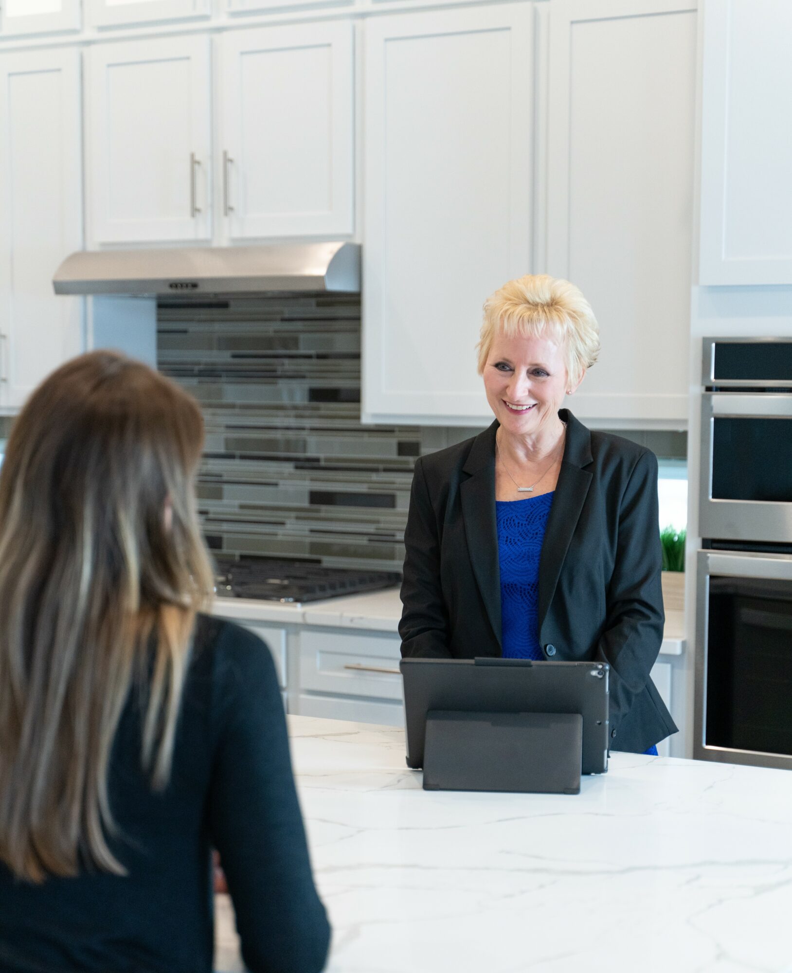 Two women talking in a modern kitchen with white cabinets and a gray backsplash, one smiling at the other.