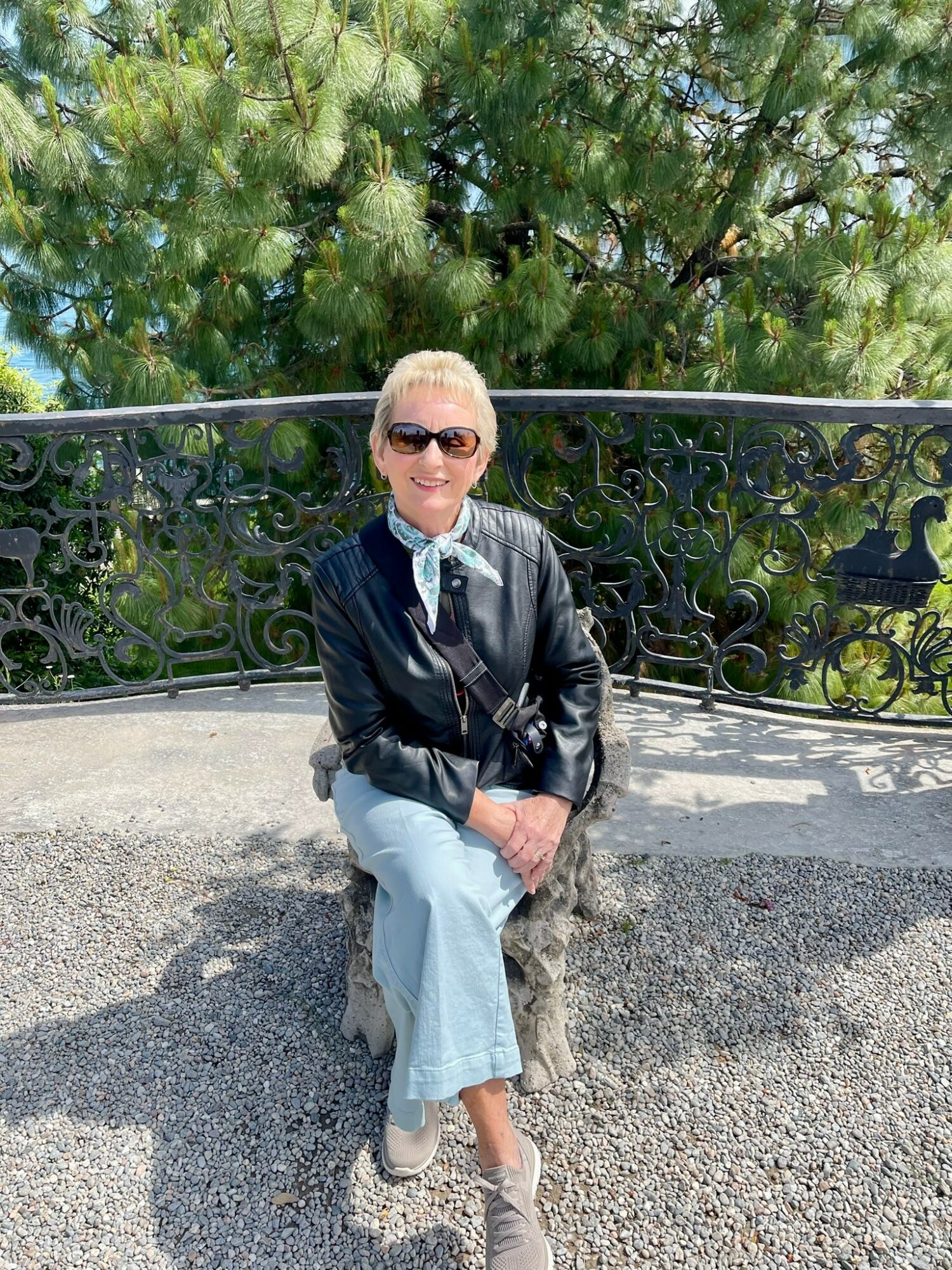 Person sitting on a stone bench outdoors, wearing sunglasses, a black jacket, and light-colored pants, with trees in the background.