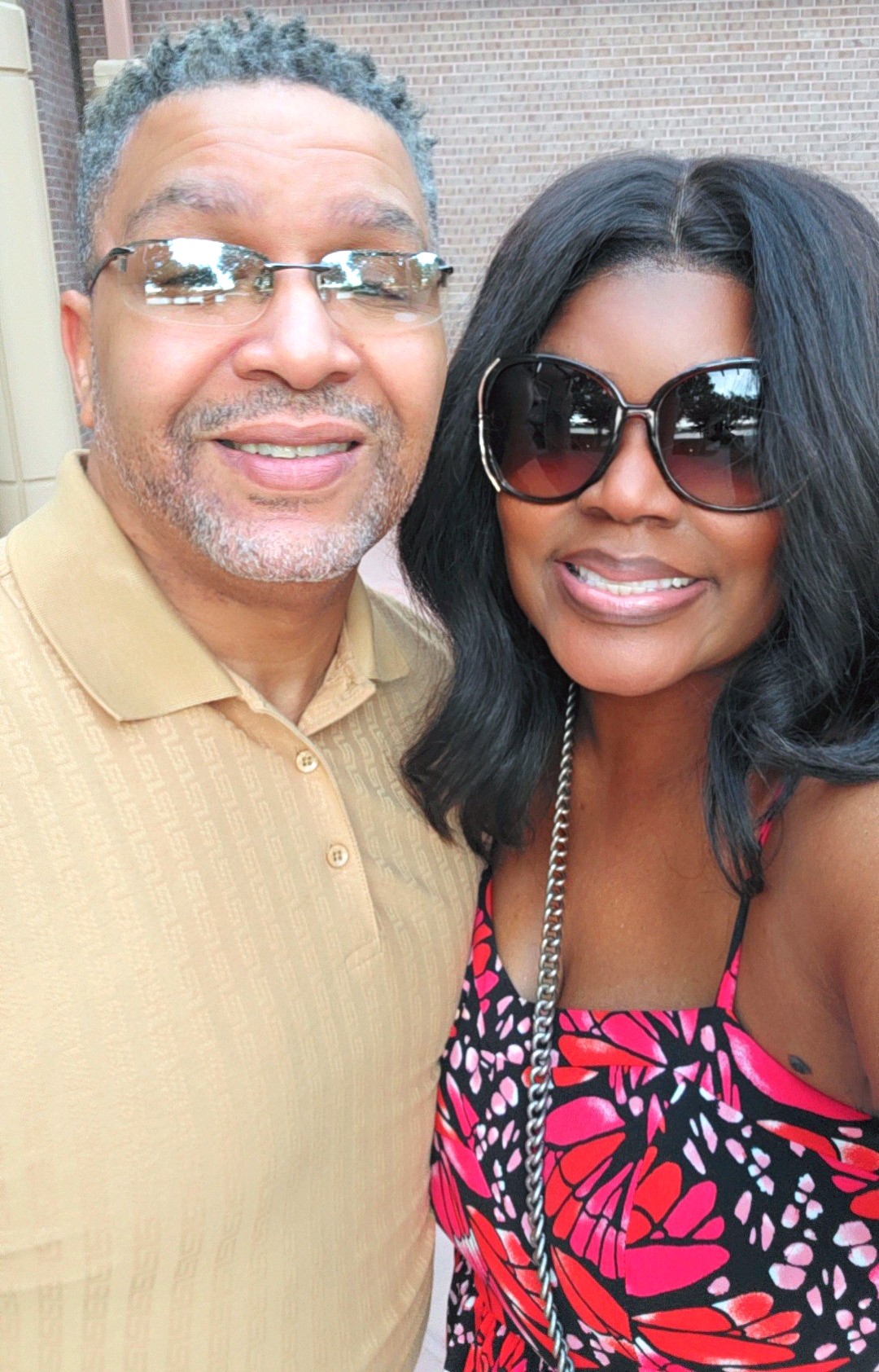 A man and woman smiling, wearing sunglasses, close together, woman in a colorful dress, man in a beige shirt.