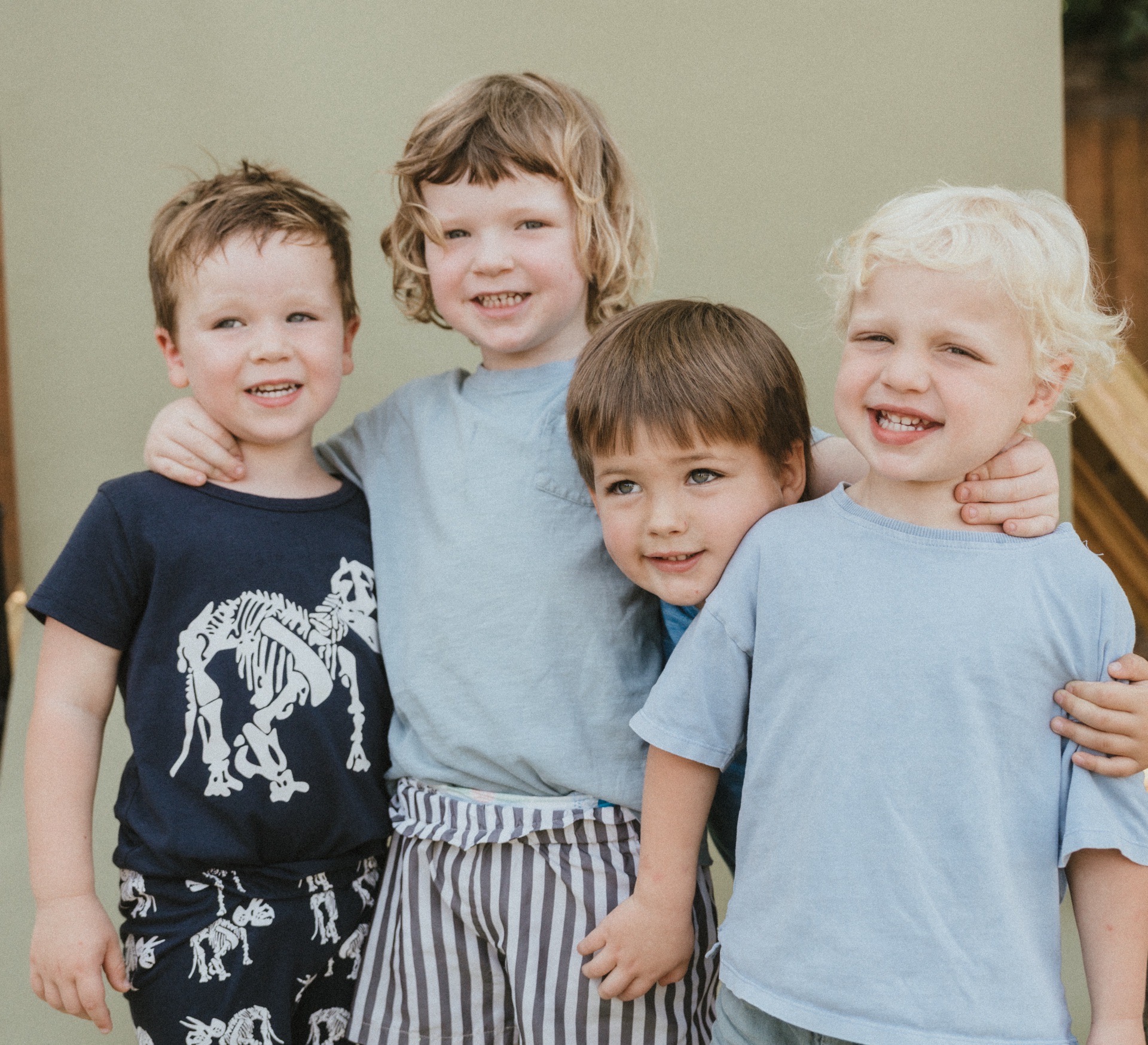 Four children smiling and standing close together outdoors, with a plain background, wearing casual clothes.