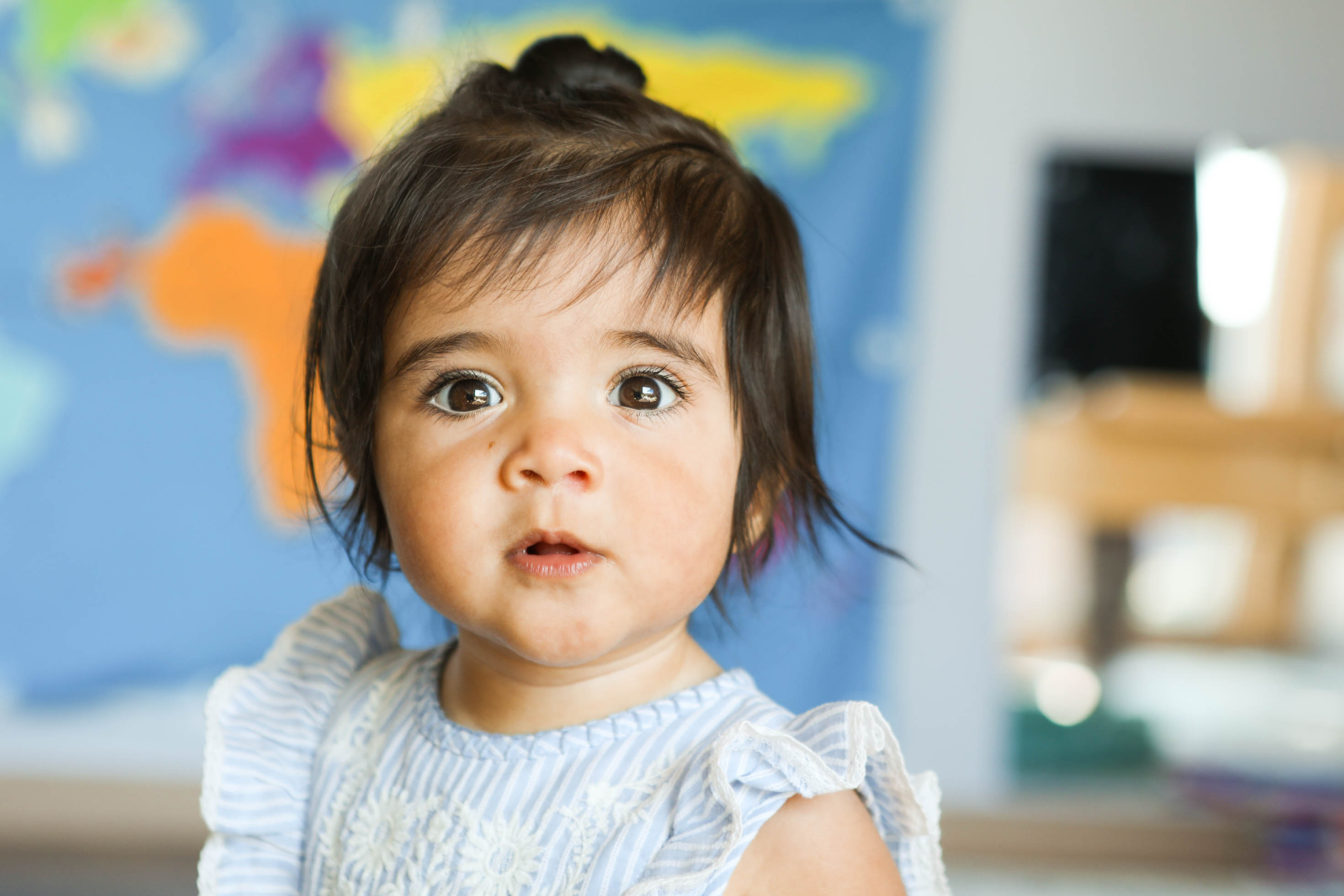 Young girl with dark hair and big eyes, wearing a light-colored top, in a room with a world map in the background.