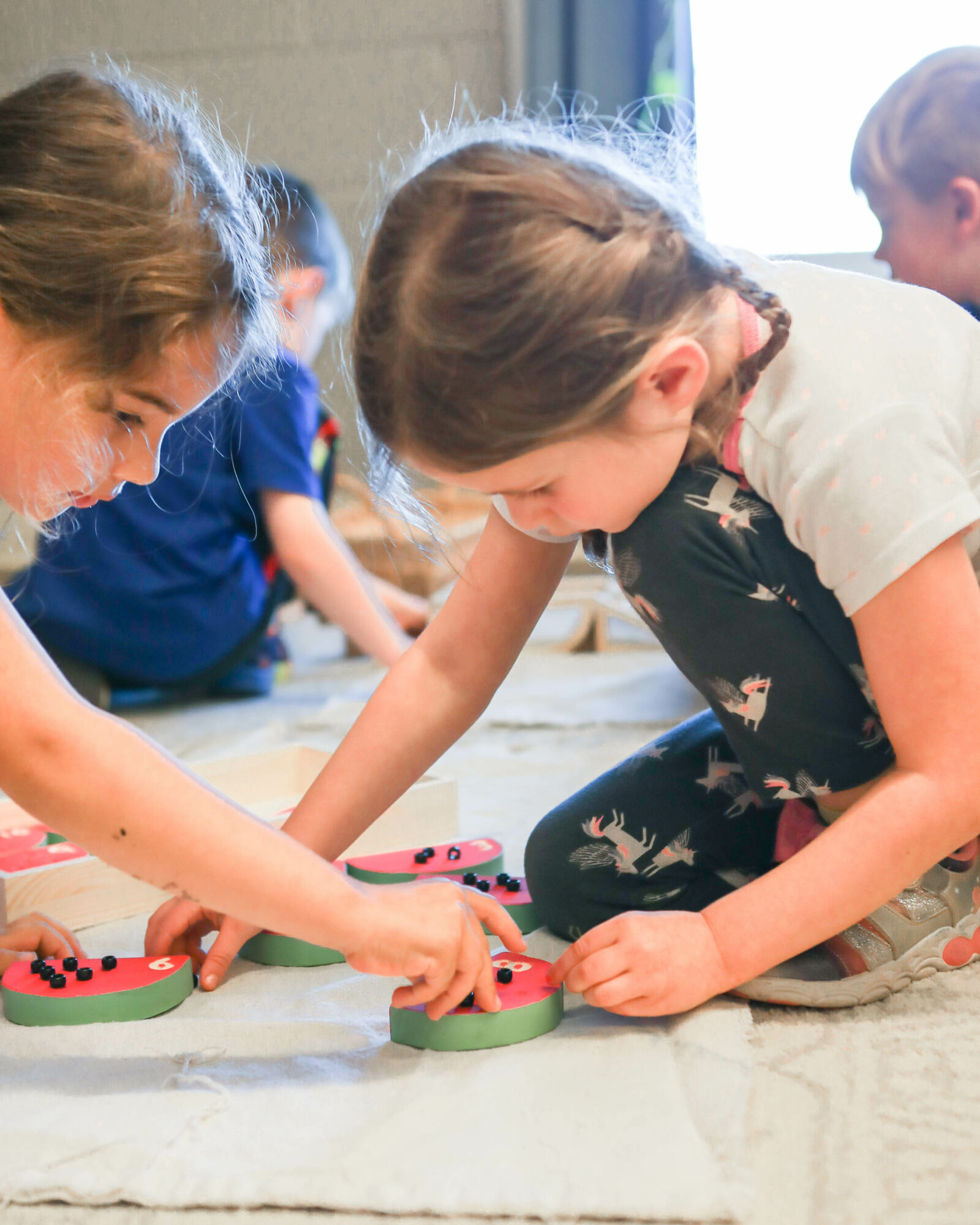 Two children are playing with colorful toy cookies on the floor, with other children in the background. Bright natural light.