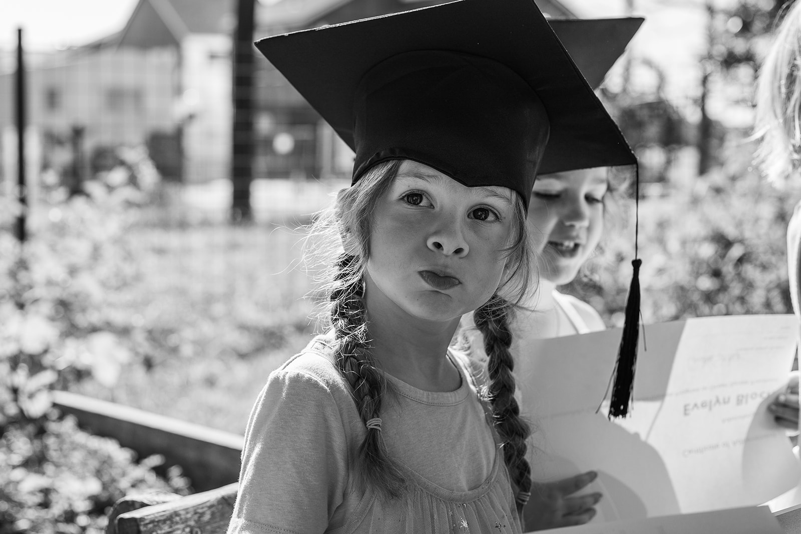 Young girl wearing a graduation cap, looking at the camera, outdoors with other children and papers nearby.
