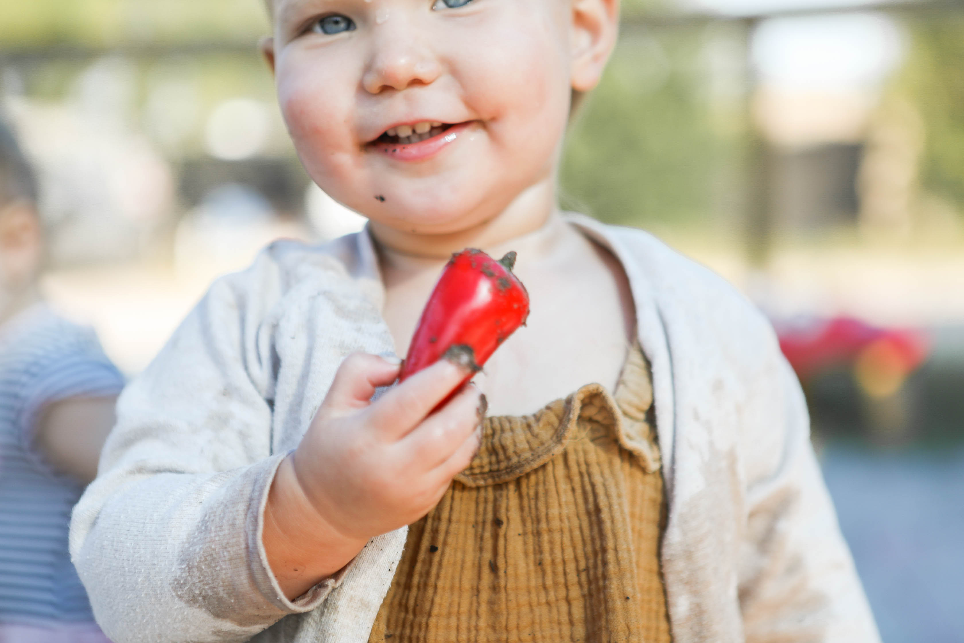 Child holding a red popsicle, smiling outdoors, wearing a brown dress and light-colored cardigan.
