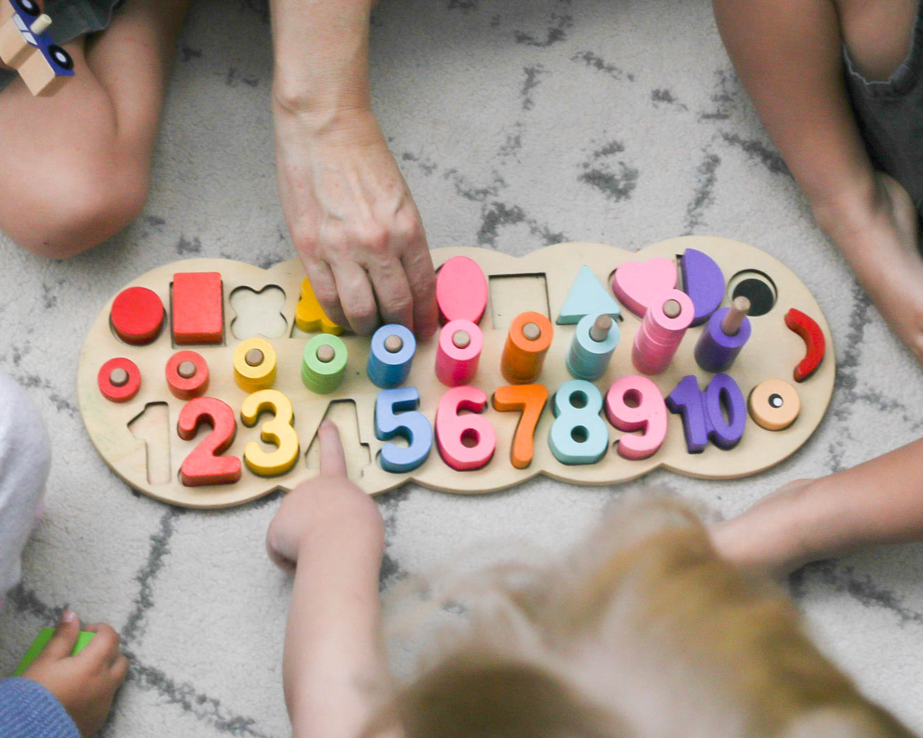 Children's hands and a dog surround a wooden number puzzle with colorful numbers and shapes, on a gray surface.