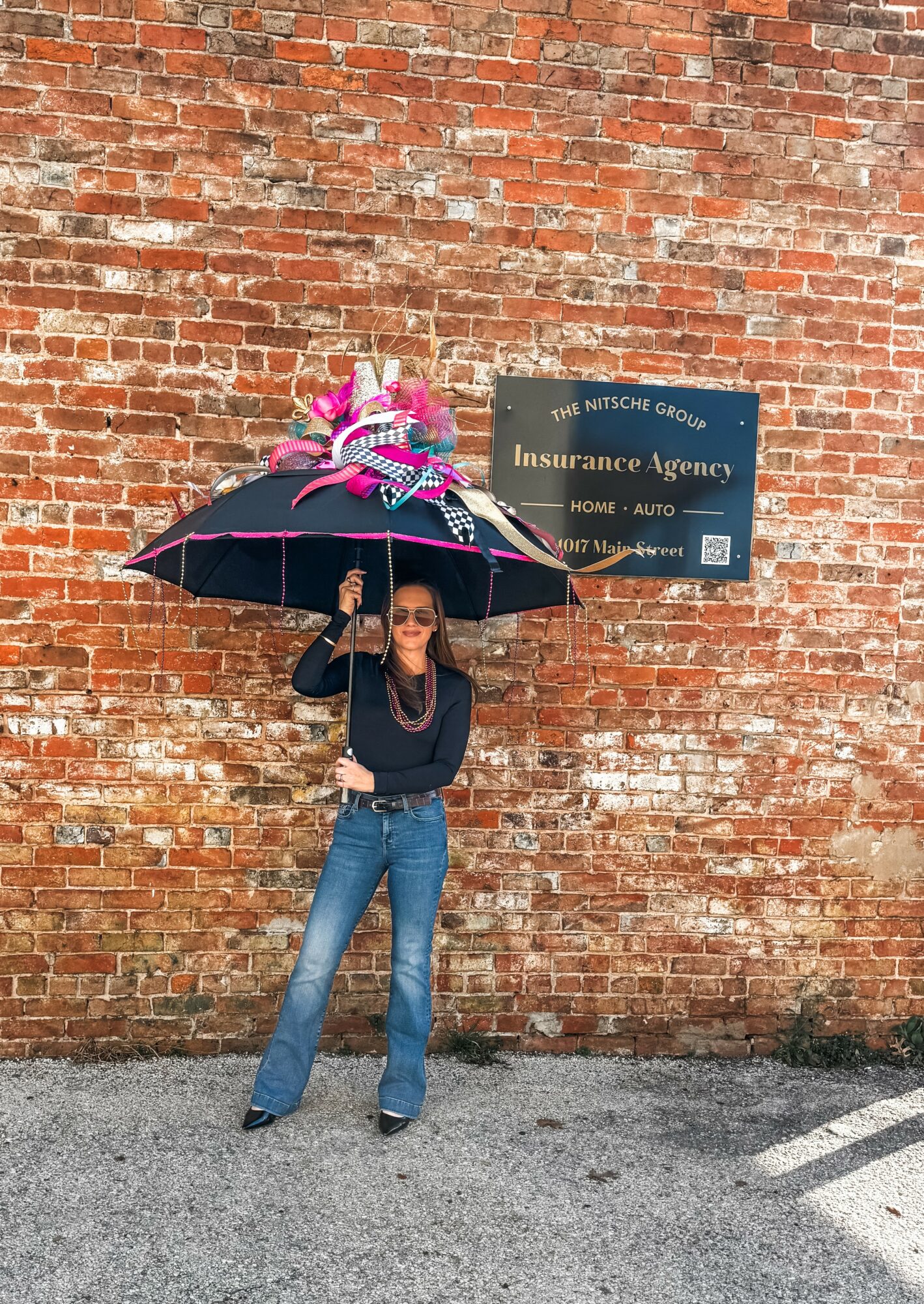 Person standing under a large umbrella with pink flowers, in front of a brick wall with a sign for an insurance agency.