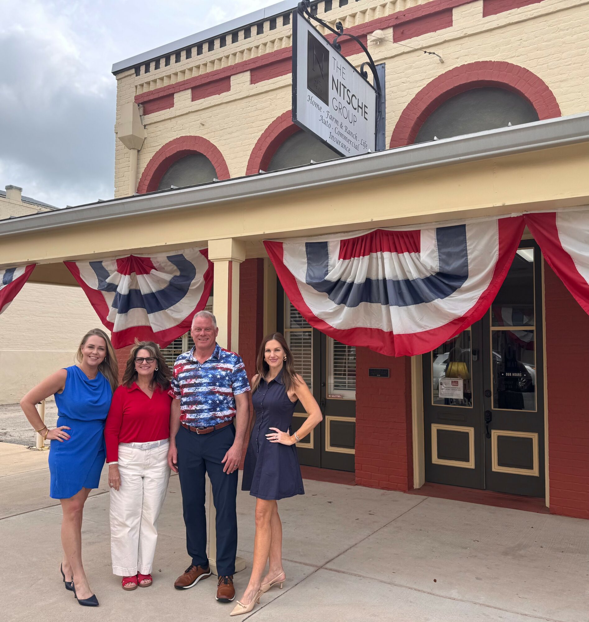 Four people standing outside a building decorated with red, white, and blue bunting, smiling at the camera.
