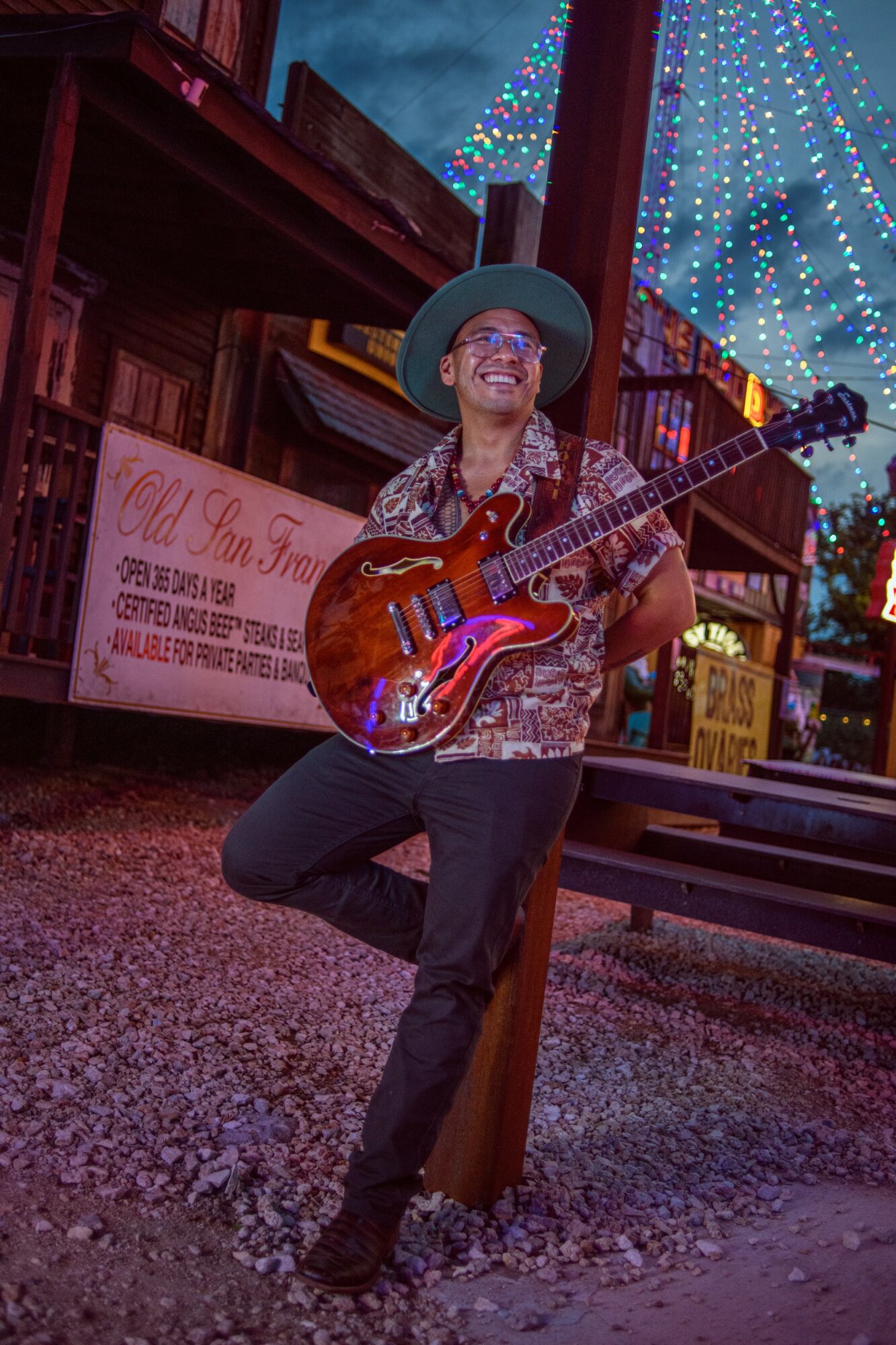 Person with a guitar, wearing a hat and sunglasses, standing outdoors near a wooden structure with colorful lights in the background.