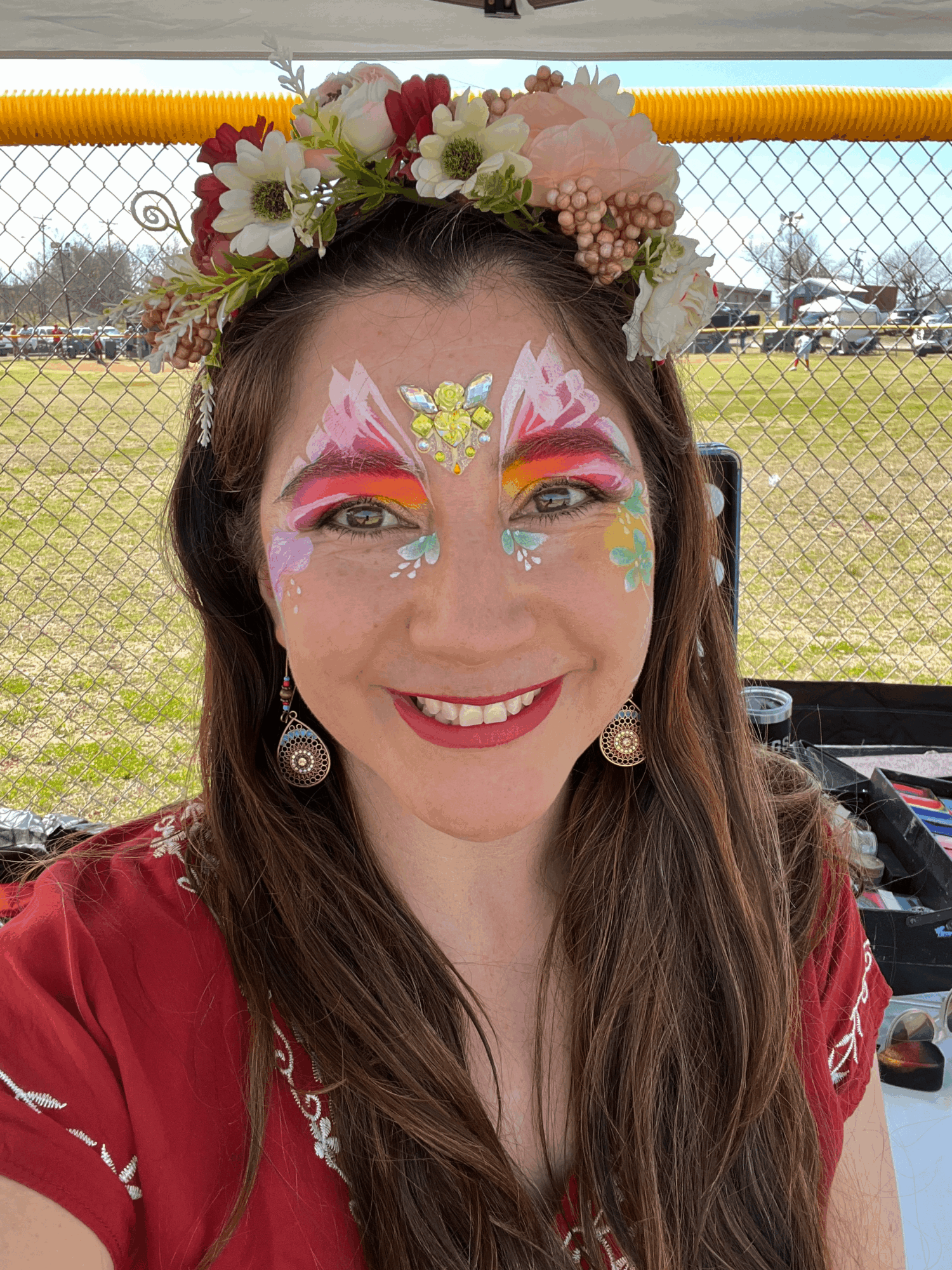 Woman with colorful face paint, floral headpiece, smiling outdoors, wearing earrings and a red top.