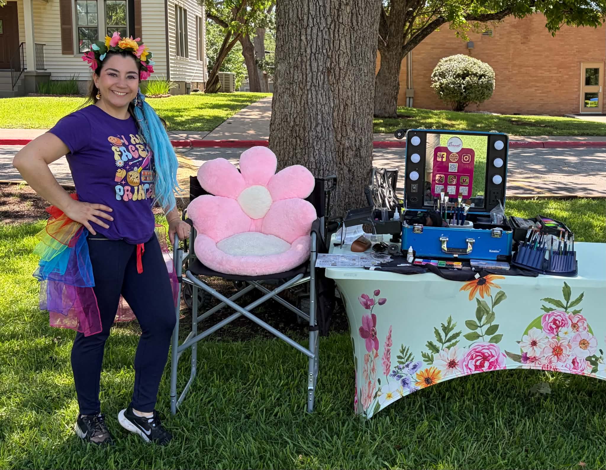 Person smiling outdoors next to a pink chair with heart-shaped cushions, a table with a floral tablecloth, and a colorful display.