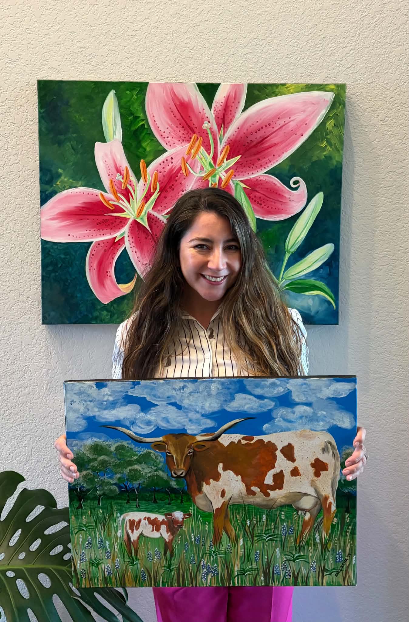 Young woman with long hair holding a painting of a cow and calf in a green field, with a blue sky background.