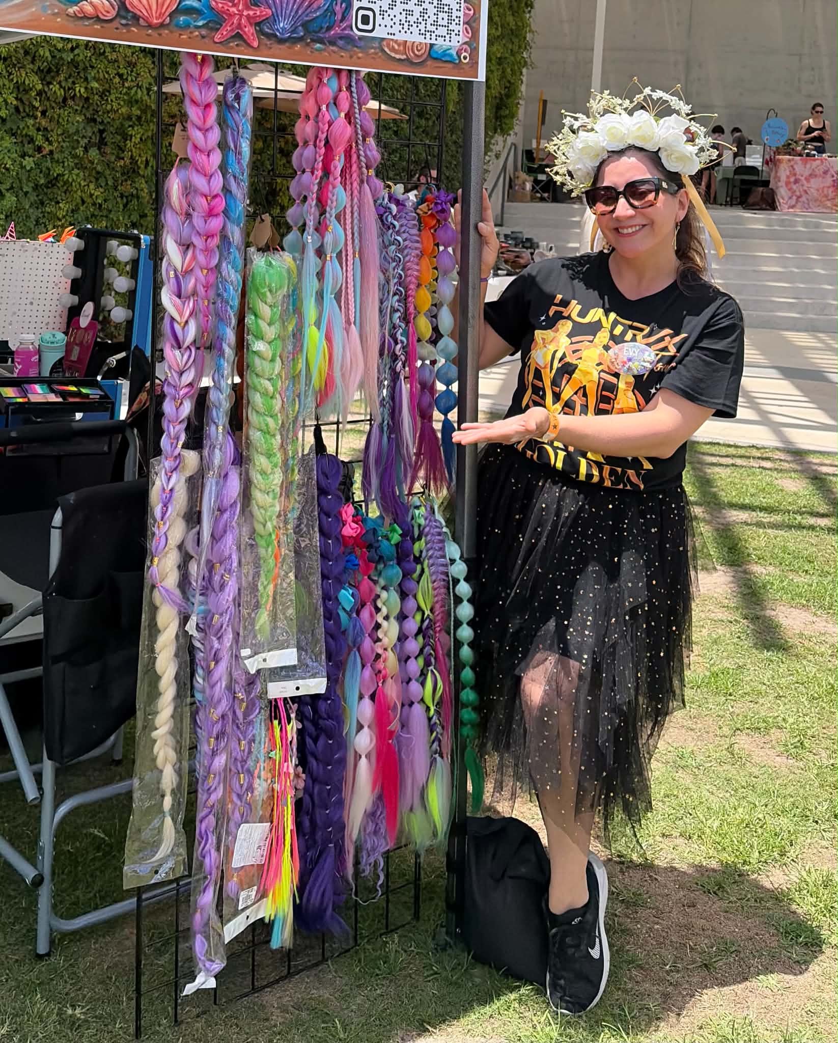 Woman wearing a flower crown and sunglasses stands next to colorful hair accessories display outdoors.