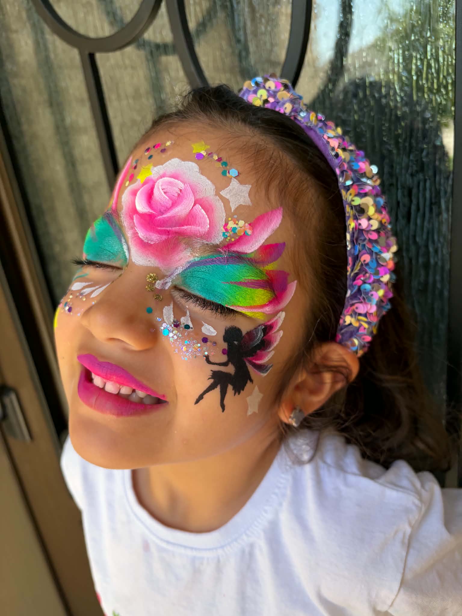 Child with colorful face paint, pink lipstick, wearing a floral headband, smiling, outdoors with a window and trees in background.