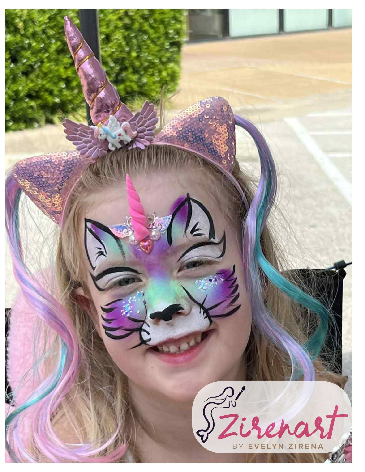 Child with colorful unicorn and cat face paint, wearing a glittery unicorn headband with ears and horn, smiling outdoors.